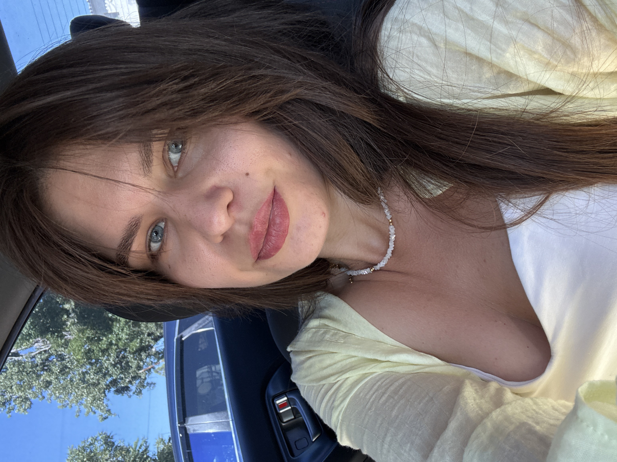 A woman with long, dark brown hair and blue eyes is seen from the chest up, looking at the camera. She is wearing a light yellow, open-front cardigan over a white top, and a beaded necklace. She is inside a car, with a car door and a car window visible behind her. Through the window, trees and a blue sky can be seen. The lighting suggests it is daytime.