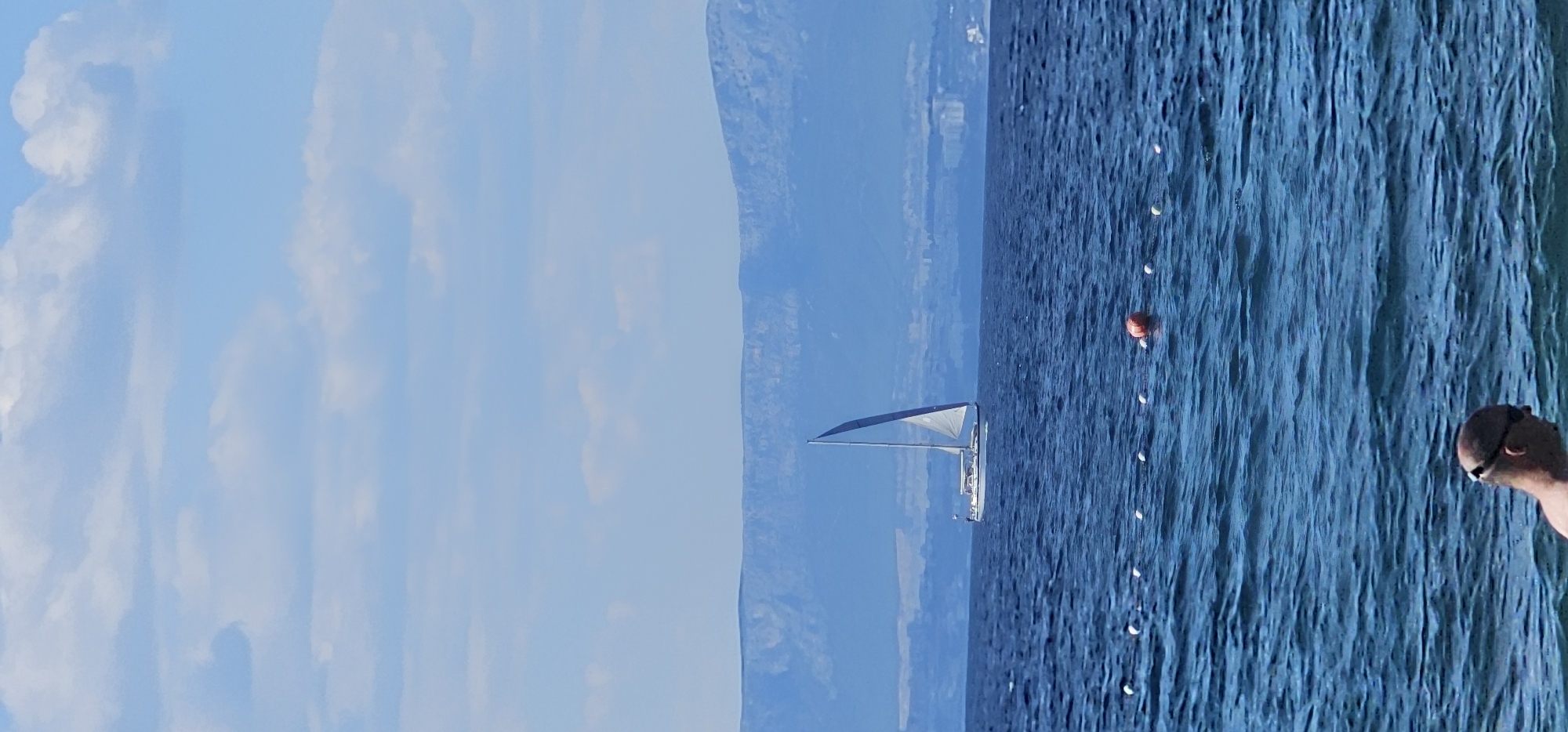 A sailboat with a white hull and a gray sail navigates the azure waters of the Adriatic Sea, with a distant mountain range forming a hazy blue backdrop. The sky above is a light blue, dotted with wispy white clouds. In the foreground, a white rope with evenly spaced white buoys stretches across the water, with an orange buoy slightly out of line with the others. The head and shoulders of a bald man wearing dark goggles are visible at the bottom of the frame, submerged in the water. The scene is likely captured from a beach in Supetar, Brač Island, Croatia, given the characteristic mountain range visible across the water, which often resembles the mainland coast visible from the island.