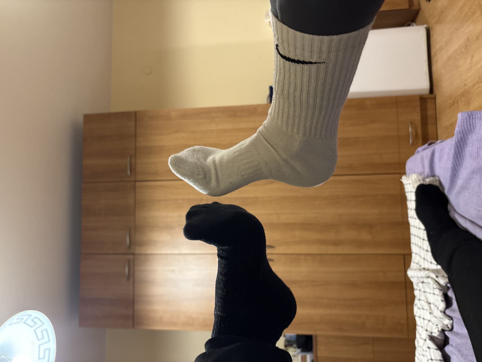 The image shows two feet wearing different colored socks, one white with a black Nike logo and one black. The feet are positioned vertically, with the white-socked foot above the black-socked foot. The background features wooden cabinets, a white rectangular object, a light fixture, and what appears to be a lavender-colored cloth draped on the floor. The location in the city of Blagoevgrad, Bulgaria is not deducible from the content of the image.