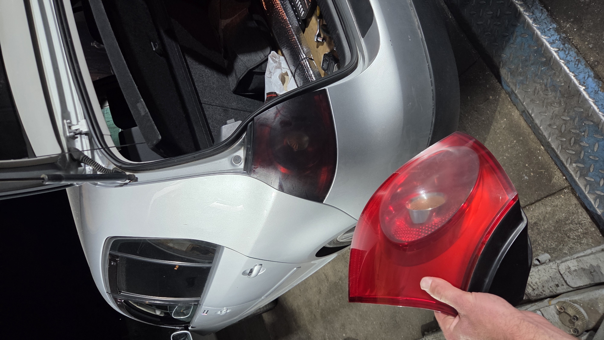 In an outdoor, dimly lit environment, a person's hand holds a red, detached taillight assembly. The taillight appears to be for a silver Volkswagen Golf hatchback. The car is parked, and its trunk is open, revealing a dark interior with some items, including what looks like a toolbox and insulation material. The car's original, darkened taillight is still attached to the vehicle's rear panel, showing a smoky or tinted finish. The ground beneath the car and the person's hand is concrete, with a metal ramp or platform visible in the foreground.