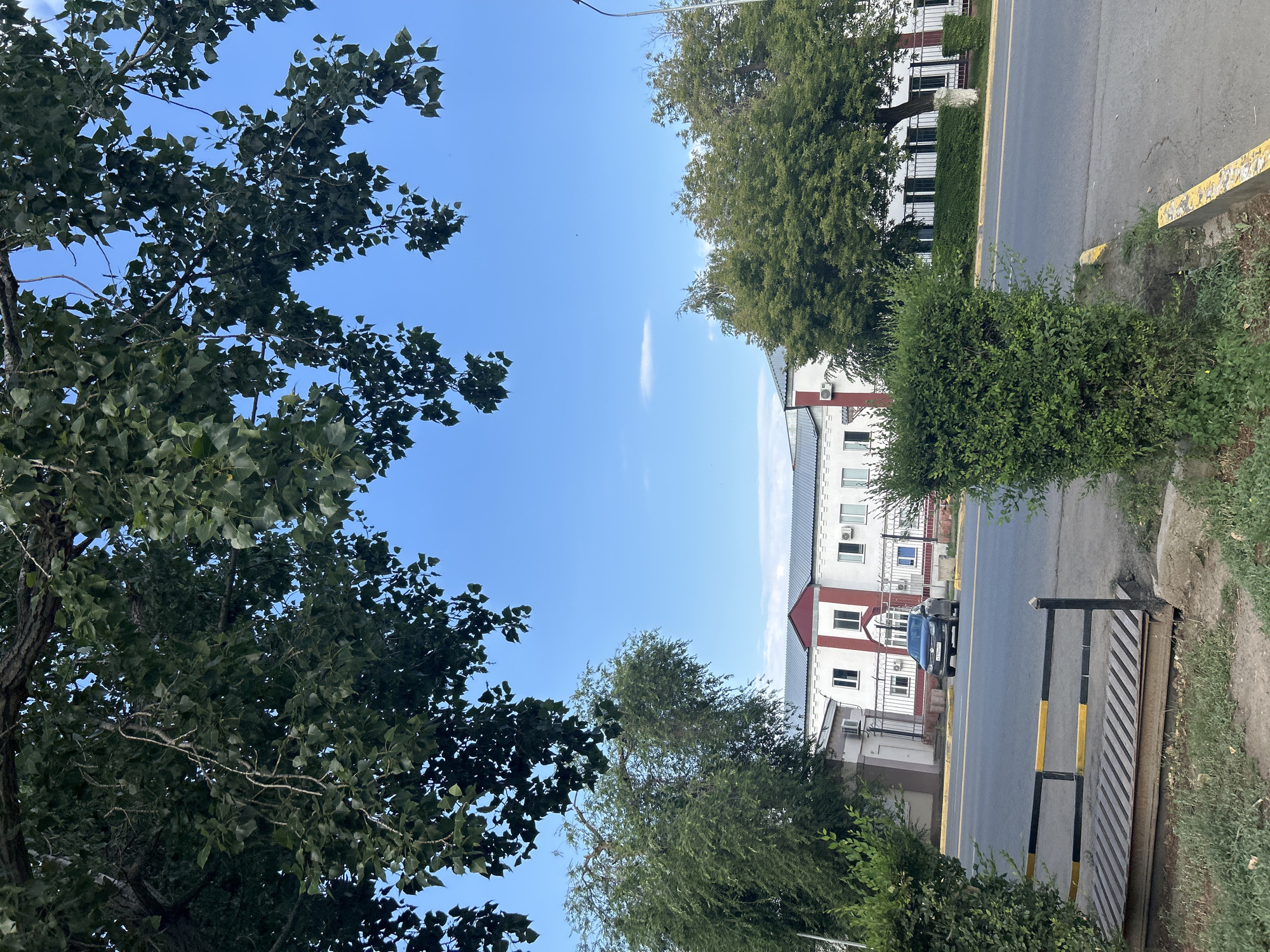 The image depicts a view of a building with white walls and a red-accented roof, situated in the city of Oral, Kazakhstan. A dark-colored sedan is parked in front of the building. The scene is bordered by lush green trees and bushes, with a street in the foreground. The sky above is clear blue with a few scattered clouds. A metal grate, likely part of a drainage system, is visible near the lower left of the frame. The overall impression is that of a quiet urban street on a pleasant day.