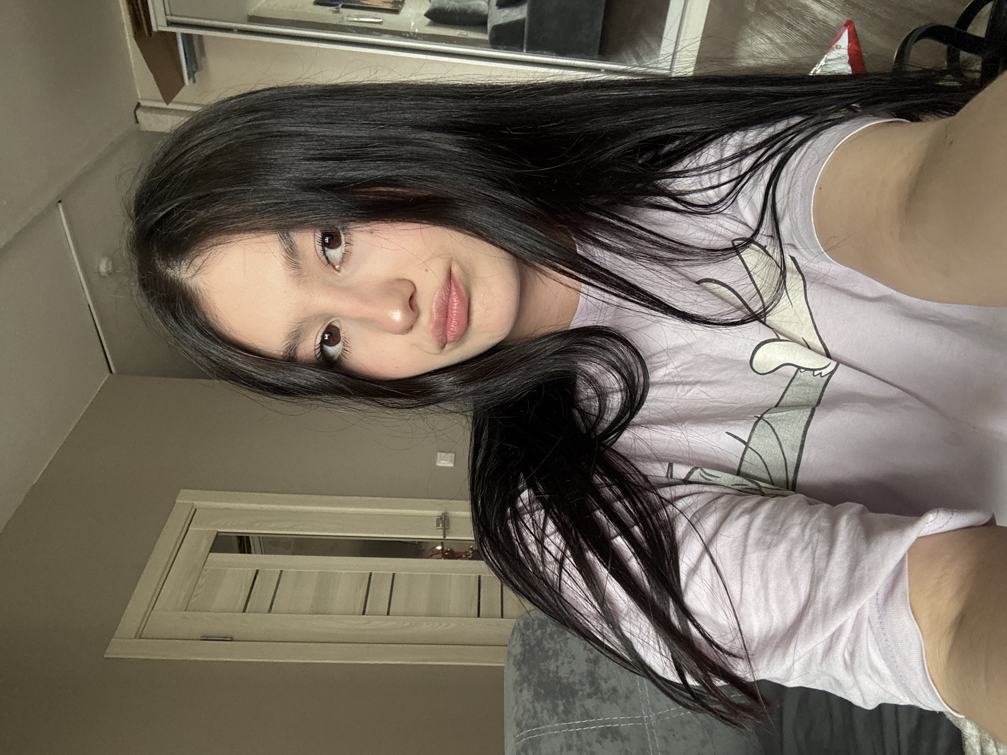The image depicts a young female, approximately 18-25 years old, in an indoor residential setting located in Chaikovsky, Russia. The female has long, straight, dark brown hair and brown eyes, looking directly at the camera with a slightly pouted expression. She is wearing a light purple short-sleeved t-shirt with a partial cartoon graphic visible. The background to her left features a grey upholstered sofa with visible stitching. Directly behind her is a neutral beige wall with a light-colored wooden door frame and a door featuring multiple horizontal panels and a frosted glass insert. To her right, a large mirrored wardrobe or closet reflects elements of the room, including a portion of the grey sofa. A white ceiling is visible at the top of the frame. Specific location details within Chaikovsky, Russia, are not discernible from the image.