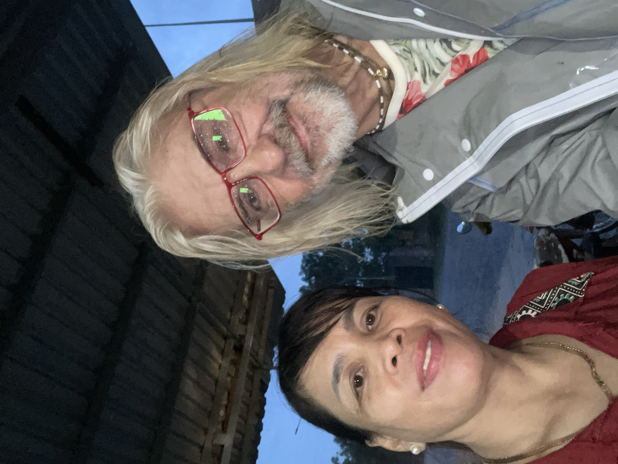 A selfie captures an older man and a woman together in an outdoor setting at dusk or early evening, possibly in Mũi Né, Vietnam.
The man, positioned on the left, has long, light blonde to white hair and a matching white beard. He wears red-framed glasses that prominently reflect green light. His attire includes a light-colored shirt with a floral pattern featuring red blossoms and green leaves, worn under a grey zipped jacket. A beaded necklace is visible around his neck. He looks towards the camera with a gentle expression.
On the right, the woman has dark hair styled with bangs. She smiles softly at the camera, wearing a reddish-brown top with a distinctive patterned collar displaying black, white, and green geometric designs. She accessorizes with small pearl earrings and a gold-colored chain necklace.
The background elements suggest an outdoor environment during twilight hours. Behind the man, a dark, slatted wooden structure is visible. To the woman's side, outlines of dark trees or foliage are present, along with a hint of a distant light against a muted blue-grey sky. The lighting is dim, creating a calm, evening ambiance. There is no visible text in the image.