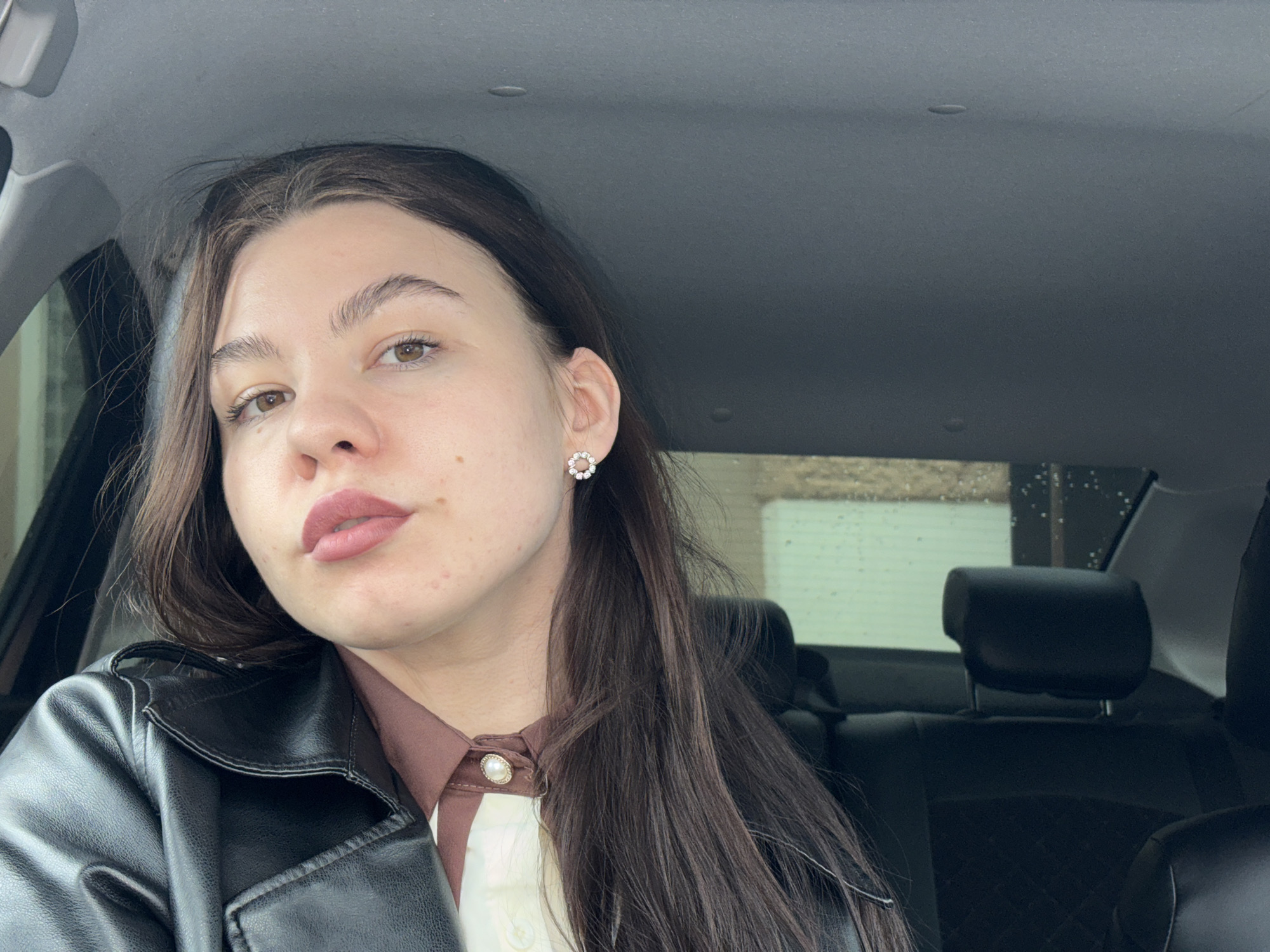 A young woman is seated in the front seat of a car. She wears a black leather jacket over a brown and white blouse with a pearl button detail. Her dark hair is parted to the side and falls over her shoulder. She has light brown eyes and is wearing a subtle lipstick. Her expression is neutral. She is wearing a small, floral-shaped earring. The background shows the interior of the car, including the headrests of the back seats. The location appears to be in the city of Perm, Russia, judging by the overcast weather and possible wet window pane visible in the background. There is no specific landmark or identifying feature that would pinpoint a precise location within Perm.