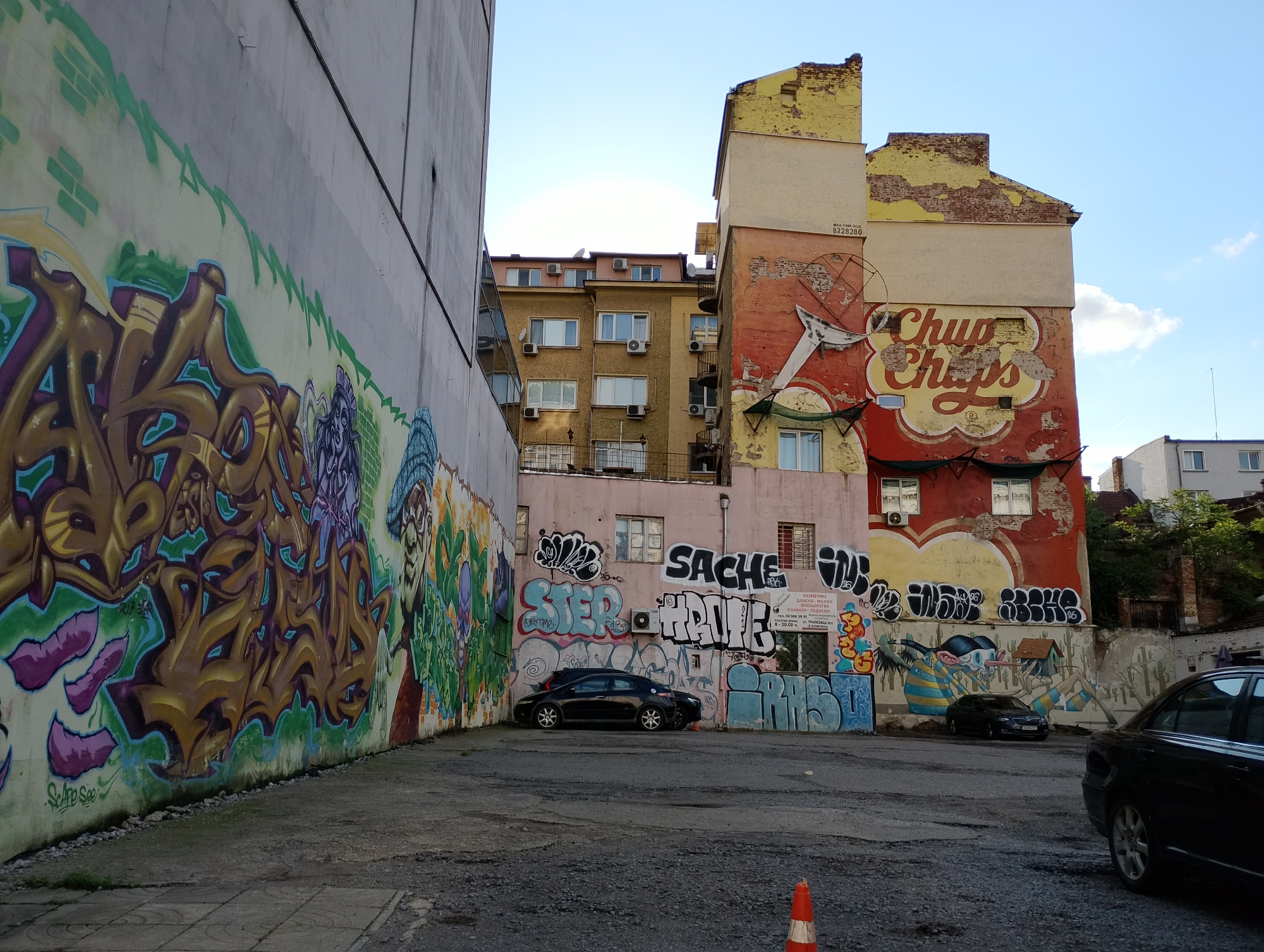 The image captures an urban alleyway or a small, informal parking area between buildings, heavily adorned with graffiti. On the left, a tall, plain wall is covered almost entirely with elaborate, colorful graffiti tags and characters, predominantly in shades of gold, purple, green, and blue. The ground in the foreground is a rough mix of asphalt and gravel, with a small orange traffic cone visible at the bottom right.
In the mid-ground, a black sedan is parked against a pink-painted building which is also covered in various graffiti tags, including "SACHE", "STER", and "HALE". A sign on this building displays Cyrillic text related to a beauty salon, including "КОЗМЕТИКА" (Cosmetics), "ДАМСКО" (Women's), "МЪЖКО" (Men's), "ФРИЗЬОРСТВО" (Hairdressing), "МАНИКЮР ПЕДИКЮР" (Manicure Pedicure), a phone number "02 986 59 02", an address "ул. ТРАПЕЗИЦА № 4", and operating hours "09.00 - 20.00 ч.".
Further back, a larger, older building with a red and yellow facade stands out. This building features a large, faded "Chupa Chups" logo painted on its side. Its upper sections show significant wear, with peeling plaster revealing brickwork. Another car, also dark-colored, is partially visible on the right. Beyond the buildings, a clear blue sky with some scattered clouds is visible, suggesting daytime, likely late afternoon due to the lighting. The overall scene depicts a vibrant yet somewhat rundown urban environment. Additional visible text includes "Scape See" and "RIP SA" on the left wall, and "МАЛ. ТИЙМ ООД" with a number "8228280" on the red building.