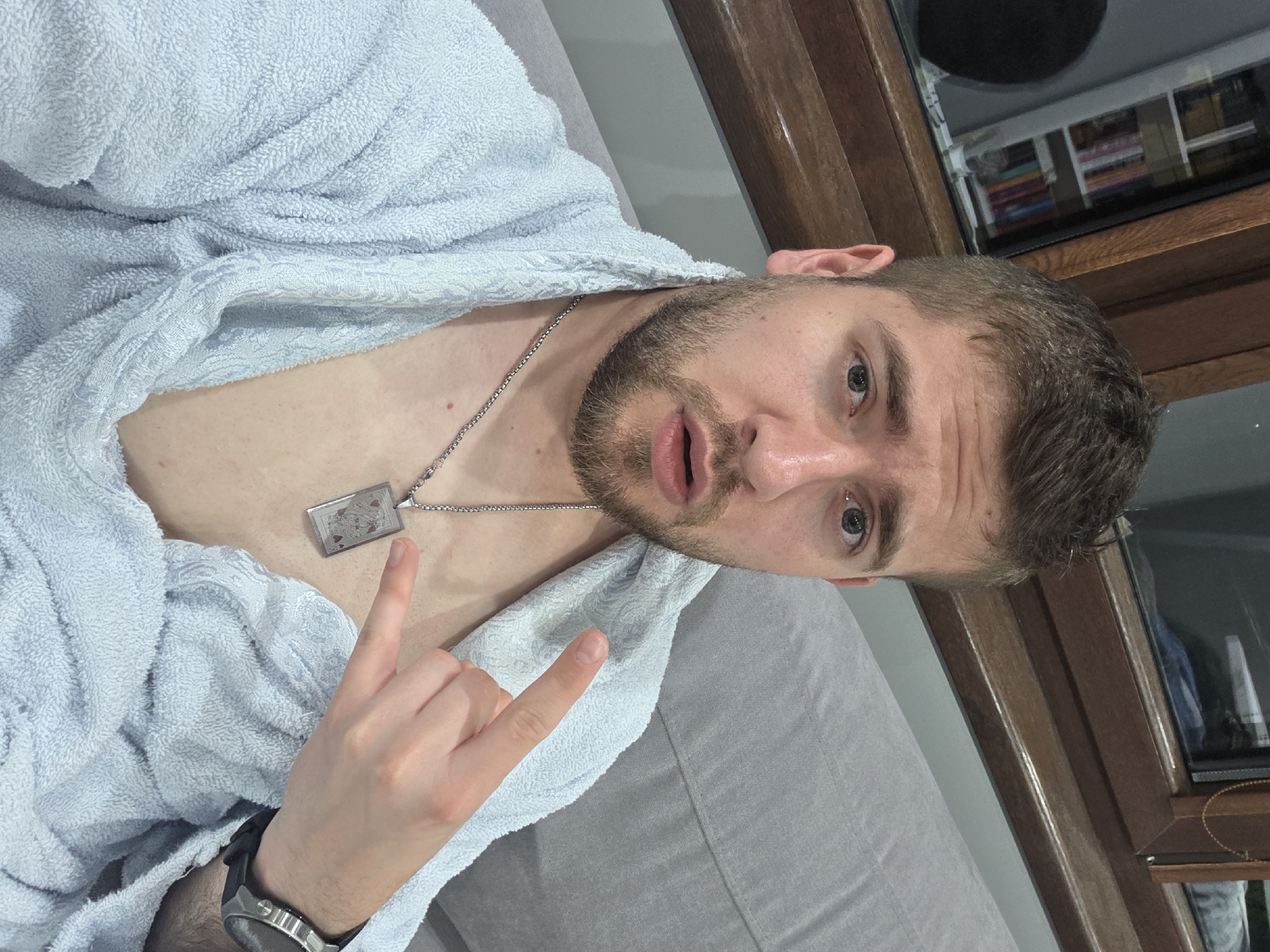 The media file presents an upward-angled selfie of a young adult male in an indoor setting. The man has light skin, short brown hair, a beard, and light eyes. He is dressed in a light blue, textured terrycloth bathrobe, which is partially open.
Around his neck, he wears a silver chain necklace with a rectangular pendant. The pendant features an intricate design resembling a playing card, likely a King or Queen of Hearts, with visible red and black accents. A metallic wristwatch with a dark strap is visible on his left wrist.
The man looks directly into the camera with a neutral expression, his mouth slightly open. His right hand is raised, performing the "rock on" hand gesture with his index and pinky fingers extended.
The background shows a gray sofa or bed where the man is reclining. To the right, a dark brown wooden frame, possibly part of a window or furniture, is prominent. Reflections in the window reveal an interior space containing shelves filled with numerous colorful items, potentially books or decorative objects. The indoor lighting provides no clear indication of the time of day. This casual scene takes place in what appears to be a residence in Yagodovo, Bulgaria. No explicit text is visible in the image, apart from the symbolic design on the necklace pendant.