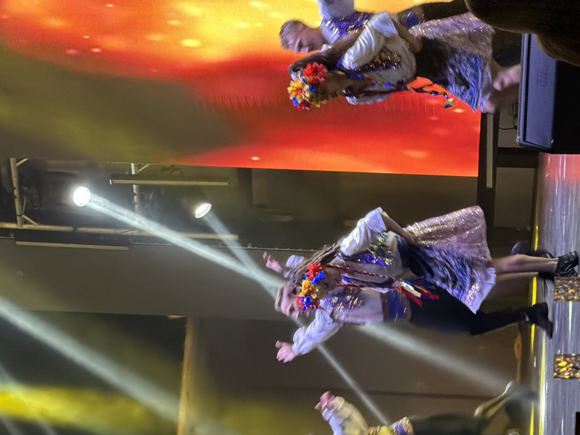 The image shows a group of performers on a stage, likely in Manavgat, Turkey, based on the provided context. There are at least four individuals visible, dressed in traditional, colorful attire adorned with sequins and floral headpieces. The performers are in motion, suggesting they are dancing. The stage is lit with bright, dramatic lighting, including yellow and white spotlights. Behind the performers, a large screen displays a vibrant gradient of orange and yellow, possibly a visual backdrop. In the foreground, there are speaker cabinets and what appear to be decorative elements on the stage floor. The overall atmosphere suggests a lively performance or cultural event.
