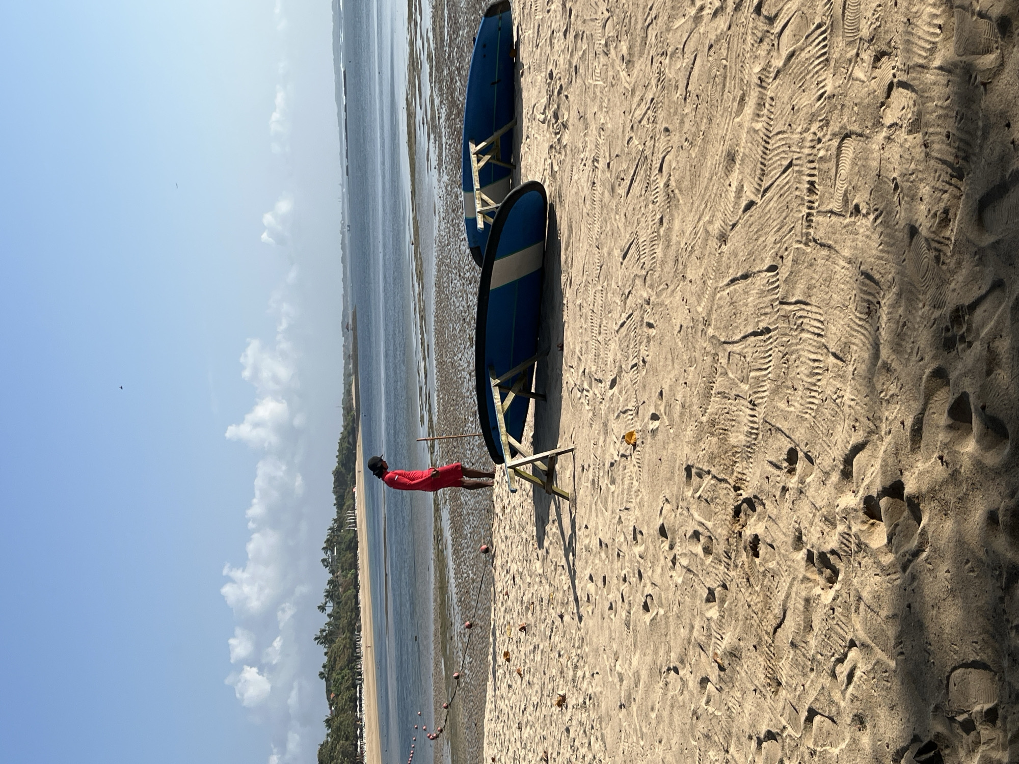 In Kuta Selatan, Indonesia, on a sandy beach, a person wearing a red long-sleeved shirt and red shorts stands near the water. The person is facing away from the camera and is looking down at the sand. Two blue surfboards are propped up on wooden stands on the beach. In the background, the ocean stretches out to the horizon under a partly cloudy sky. The sand is covered in footprints.