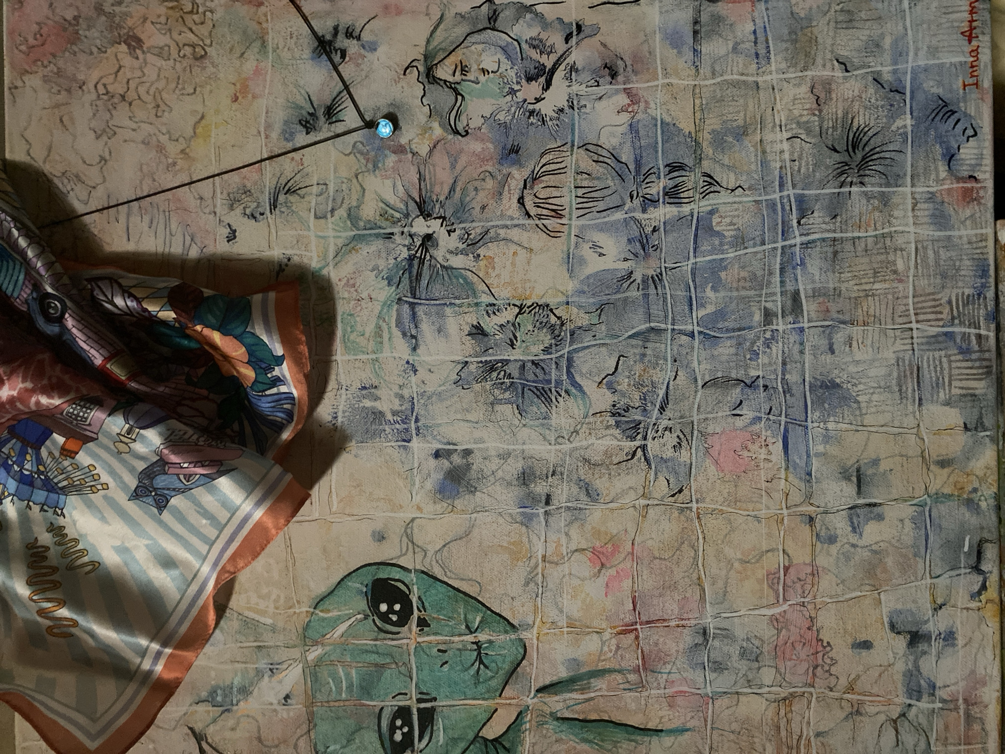 The image is a close-up shot of a mixed-media artwork. The artwork appears to be on a textured canvas, with elements of painting, drawing, and fabric collage. In the upper left corner, a brightly colored silk scarf is draped over the canvas. The scarf has a pattern with geometric shapes, wavy lines, and what appear to be stylized buildings or abstract forms in blues, whites, oranges, and pinks.
The canvas itself is covered with a background of abstract washes in earthy tones, pastel pinks, and blues. Overlaid on this background are various artistic elements. On the left side of the canvas, a stylized green alien head with large black eyes is depicted. The alien is rendered in a somewhat cartoonish, graphic style.
Further to the right, there are charcoal or ink drawings of human faces and floral or abstract organic shapes. These drawings are layered on top of the painted background and some appear to be partially obscured by the fabric of the scarf. A thin black wire or string is visible extending from the upper right portion of the canvas downwards, with a small, glowing blue light attached to it. This light could be an LED or a reflective element.
The entire canvas is overlaid with a white grid pattern, which gives the impression of a window pane or a screen, dividing the artwork into segments. The texture of the canvas is visible through the grid. In the bottom right corner of the artwork, the signature "Inna Arm..." is visible in red script.
The artwork is displayed indoors, as suggested by the lighting and the presence of the scarf and wire. There are no elements in the image that definitively place it in Thira Municipal Unit, Greece.