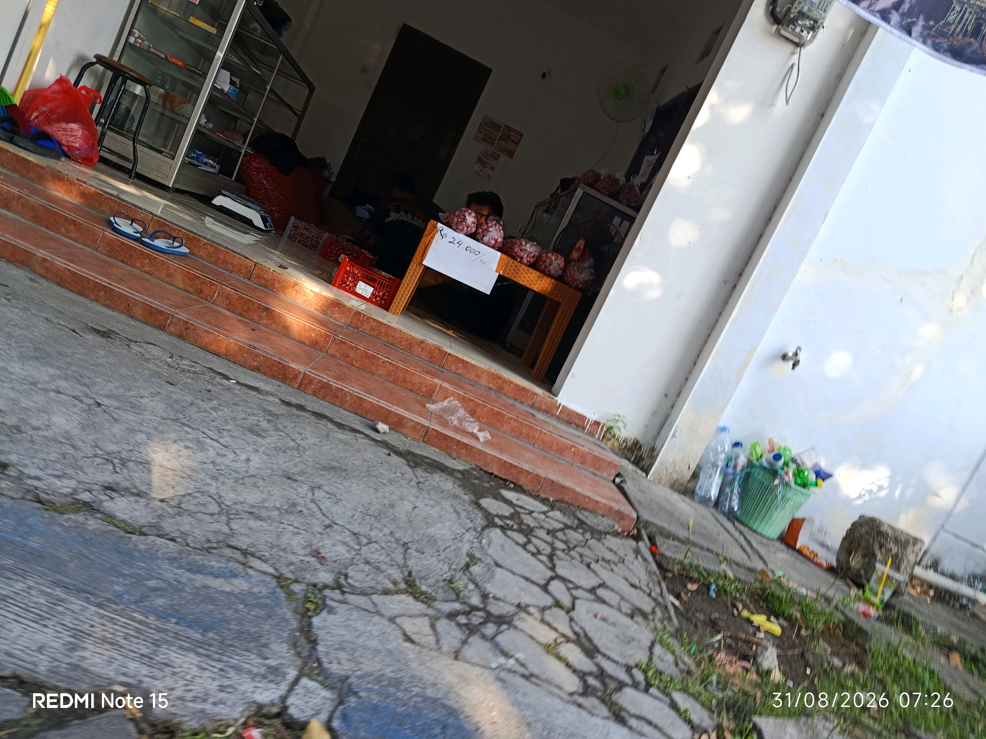 The image captures an outdoor scene in front of a shop, likely in Keboansikep, Indonesia, as suggested by the timestamp and potential local context. The foreground is dominated by cracked concrete paving stones with patches of grass growing through them. A set of reddish-brown tiled stairs leads up to the shop entrance. On the stairs, a pair of blue and white flip-flops are casually placed.
The shop itself appears to be a small retail establishment. Inside, a display cabinet with glass shelves is visible, containing various items. To the right of the entrance, a small table is set up, displaying several bags of red onions. A white sign on the table reads "Rp 24.000/Kg", indicating the price of the onions. A person is partially visible behind the table, tending to the produce.
Further to the right, near the wall of the building, is a green basket filled with plastic bottles and other assorted items, suggesting it might be a collection point for recyclables or general refuse. A large rock is also placed near the basket. The building's exterior wall is painted white. The overall impression is of a humble, everyday street scene at a local shop. The timestamp "31/08/2026 07:26" is superimposed on the image.