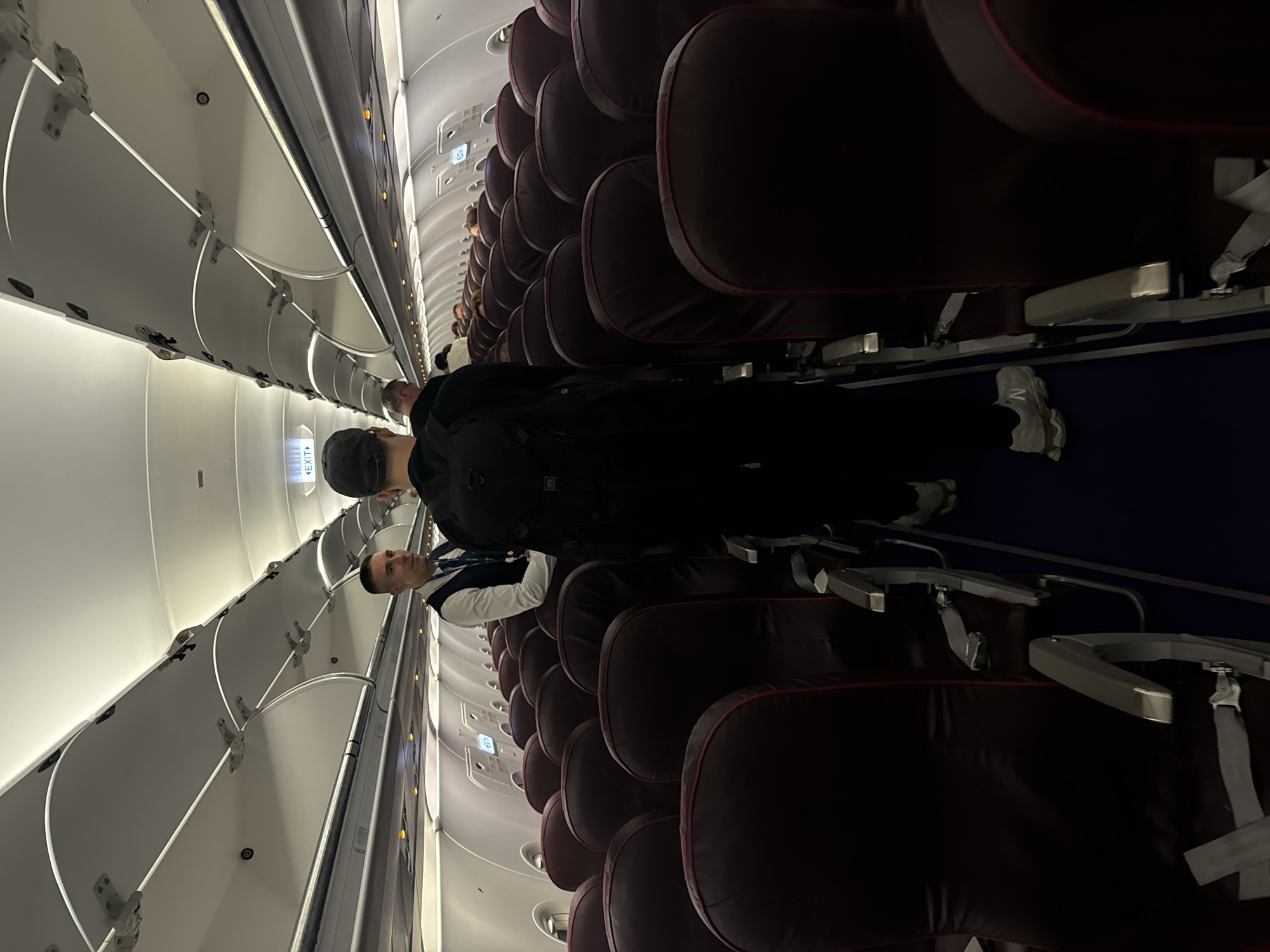 The image depicts the interior of an airplane cabin. Passengers are standing in the aisle. Rows of seats are visible on both sides of the aisle. The seats are upholstered in a dark color. The overhead compartments are visible above the seats. A flight attendant is standing in the aisle. The image was taken in an aircraft, not in Khimki, Russia.