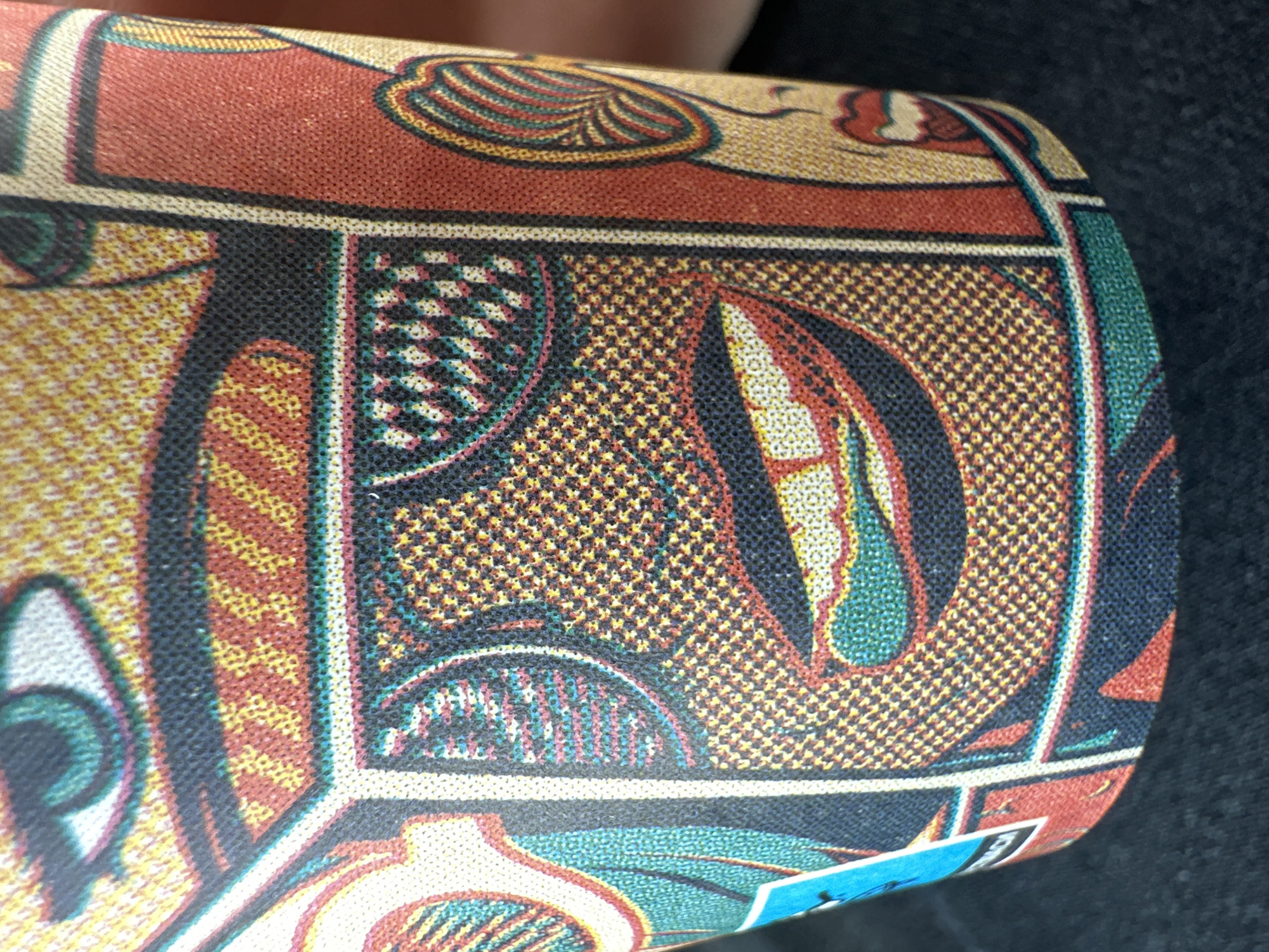 The image shows a close-up of patterned fabric wrapped around a cylindrical object. The fabric features geometric and stylized organic shapes in a palette of red, yellow, blue, black, and white. The pattern is bordered by darker lines. A small piece of a blue tag is visible at the bottom right corner. The background is a dark, out-of-focus texture that appears to be another surface. The image provides no information related to the city of Dolna Malina, Bulgaria.
