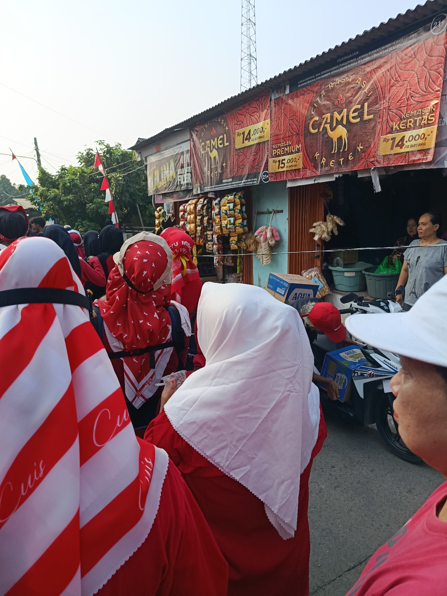 This is a medium-shot, eye-level, outdoor, medium-distance, horizontal photograph taken in Balaraja, Indonesia. The scene depicts a street lined with shops and adorned with Indonesian flags. A group of people, many wearing red and white attire, are gathered. One individual in the foreground wears a white headscarf and a red shirt, with a portion of another person's pink shirt visible. Another person on the left is wearing a white and red striped headscarf. A motorcycle with an "AQUA" box on it is parked in the middle ground, and behind it, a group of women are standing outside a shop. A large banner advertising "CAMEL KERTAS" cigarettes hangs above the shop, featuring prices and warnings. The overall atmosphere appears to be that of a local gathering or event.