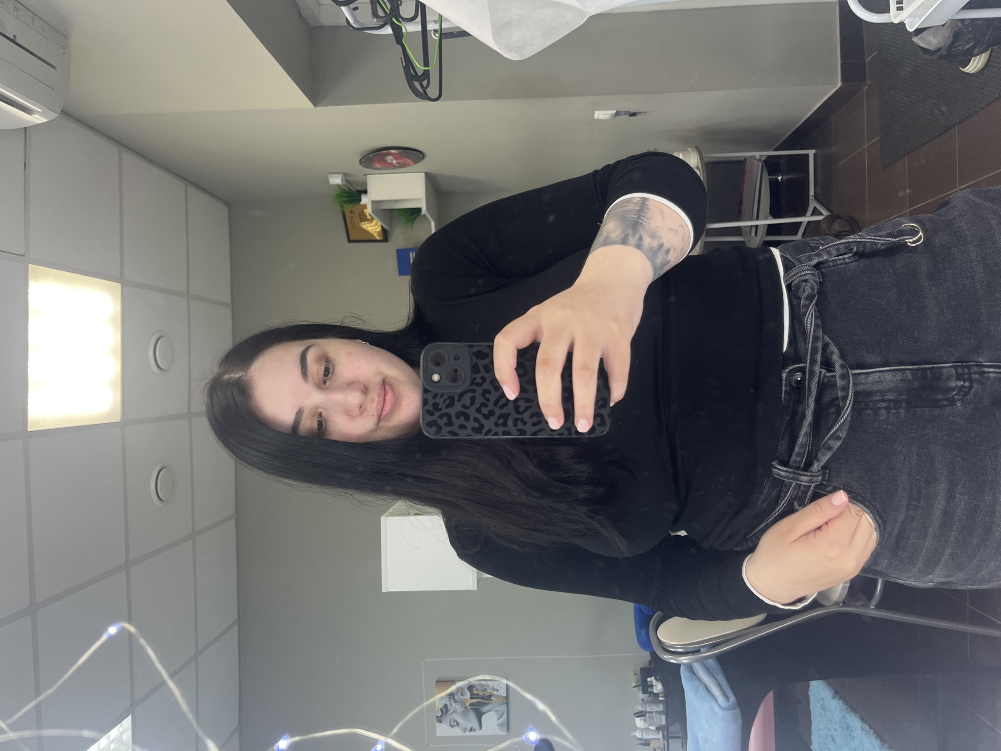 A young woman with long, dark hair is captured in a mirror selfie, standing in what appears to be an indoor commercial or studio setting, possibly in Chaikovsky, Russia. She wears a black long-sleeved top and black high-waisted jeans with a white drawstring detail, and her fingernails are painted a light pink. Her left forearm displays a distinctive tattoo depicting a forest or tree landscape in grey tones. She holds a black smartphone with a leopard-print case up, its camera facing the mirror to capture her reflection. A slight smile is visible on her face, suggesting a neutral or content mood.
The background, reflected in the mirror, shows a well-lit room with a modern aesthetic. The ceiling features white grid tiles with both rectangular and circular recessed lights. An air conditioning unit is visible high on the wall to her right, above a section of white cabinets or shelving. Further right, a small green potted plant and a framed item are on the wall. To her left, a white chair with a metallic frame and a blue cushion is seen, along with a framed artwork of a classical sculpture's head. Delicate string lights with blue-ish bulbs are draped across the lower part of the mirror's surface. The floor appears to be tiled in a dark color, and a dark mat is also visible. No discernible text is visible in the image. The setting suggests artificial lighting, providing no cues about the time of day or outdoor weather.