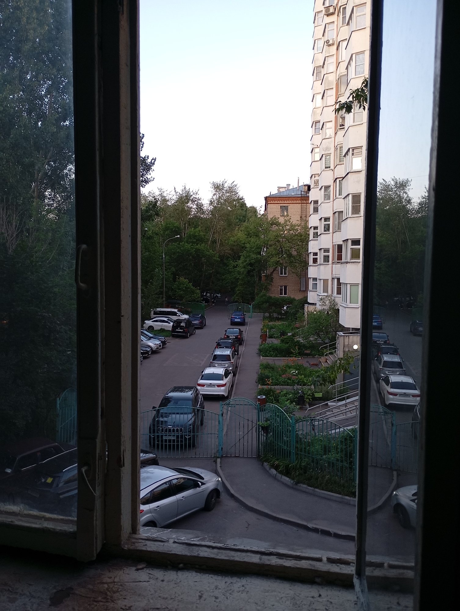 The image captures a view from a window overlooking a residential courtyard in Moscow, Russia. The scene is characterized by a multi-story apartment building to the right, its facade a neutral light color with numerous windows. In front of the building, a parking lot is visible, filled with various cars. Some of these cars are parked along the side of the building, while others are arranged on a paved road that curves through the courtyard.
To the left of the apartment building, another, older brick building is partially visible, surrounded by trees. The courtyard is interspersed with green spaces, including planted areas with shrubs and trees that line the walkways and parking areas. A distinctive green metal gate is visible, suggesting an entrance or partition within the courtyard. The sky above is a pale, clear blue, indicating it might be late afternoon or early evening. The foreground of the image shows the interior of the room, with a wooden window frame and sill, suggesting the photo was taken from inside an apartment. The overall impression is a typical urban residential setting.