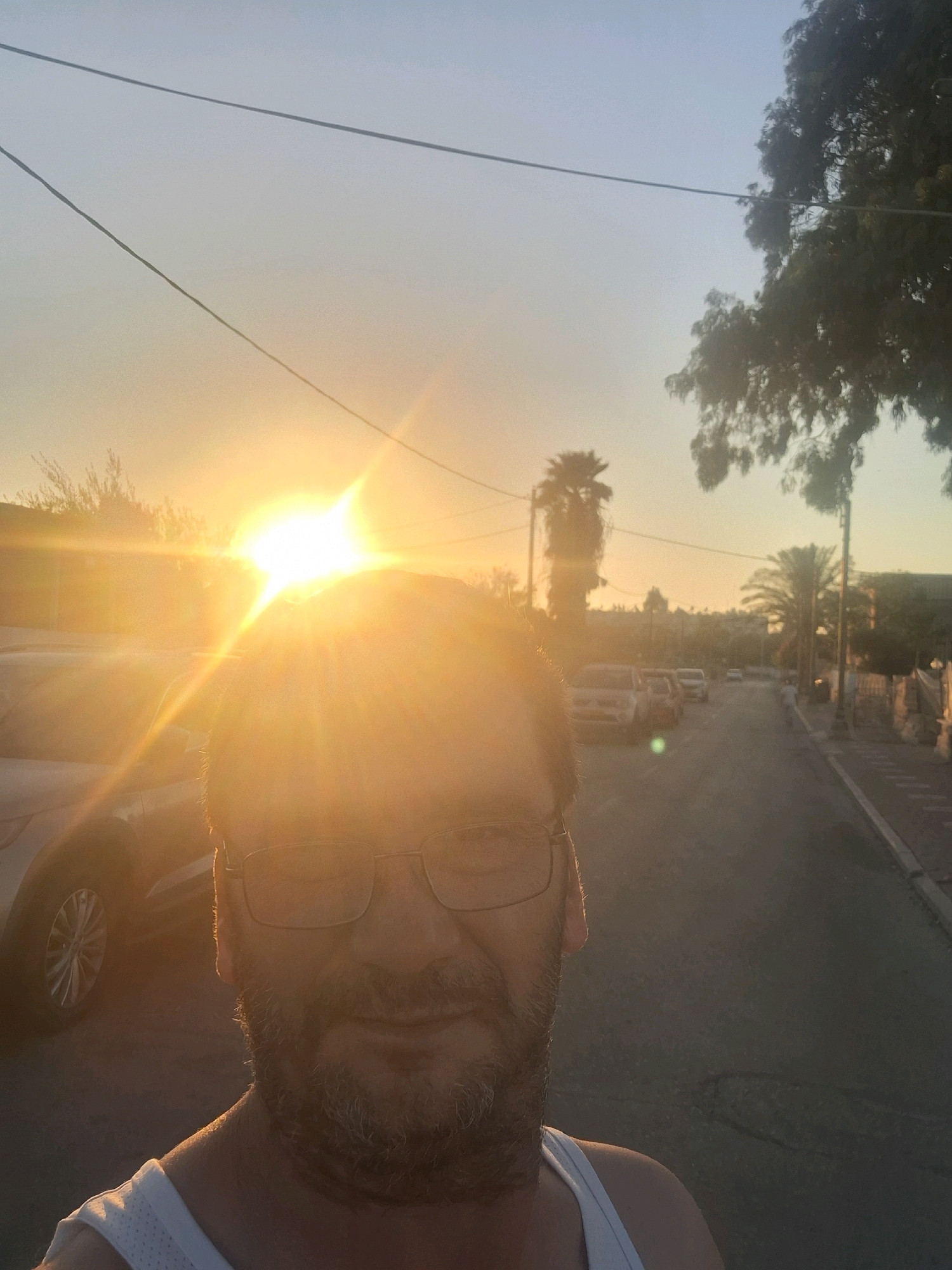 A man is taking a selfie on a street in Yerocham, Israel, during sunset. The sun is very bright, causing lens flare and obscuring some details of the background. The man is wearing glasses and a white tank top. To his left, a white SUV is parked. The street is paved, and there are cars parked on both sides. Palm trees and buildings are visible in the distance. Power lines are strung across the sky. The overall atmosphere suggests a warm, late afternoon in a residential area.