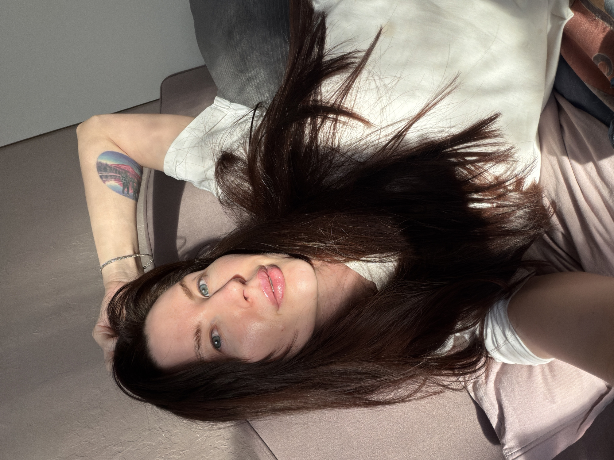 A woman with long, dark brown hair lies on a mauve-colored sofa. She is wearing a white T-shirt. Her left arm is raised, revealing a tattoo on her forearm and a silver bracelet. The woman's face is visible, showing light skin, blue eyes, and full, glossy lips. Light is streaming in, creating shadows on the wall behind the sofa. The location of Perm, Russia cannot be determined from the image.