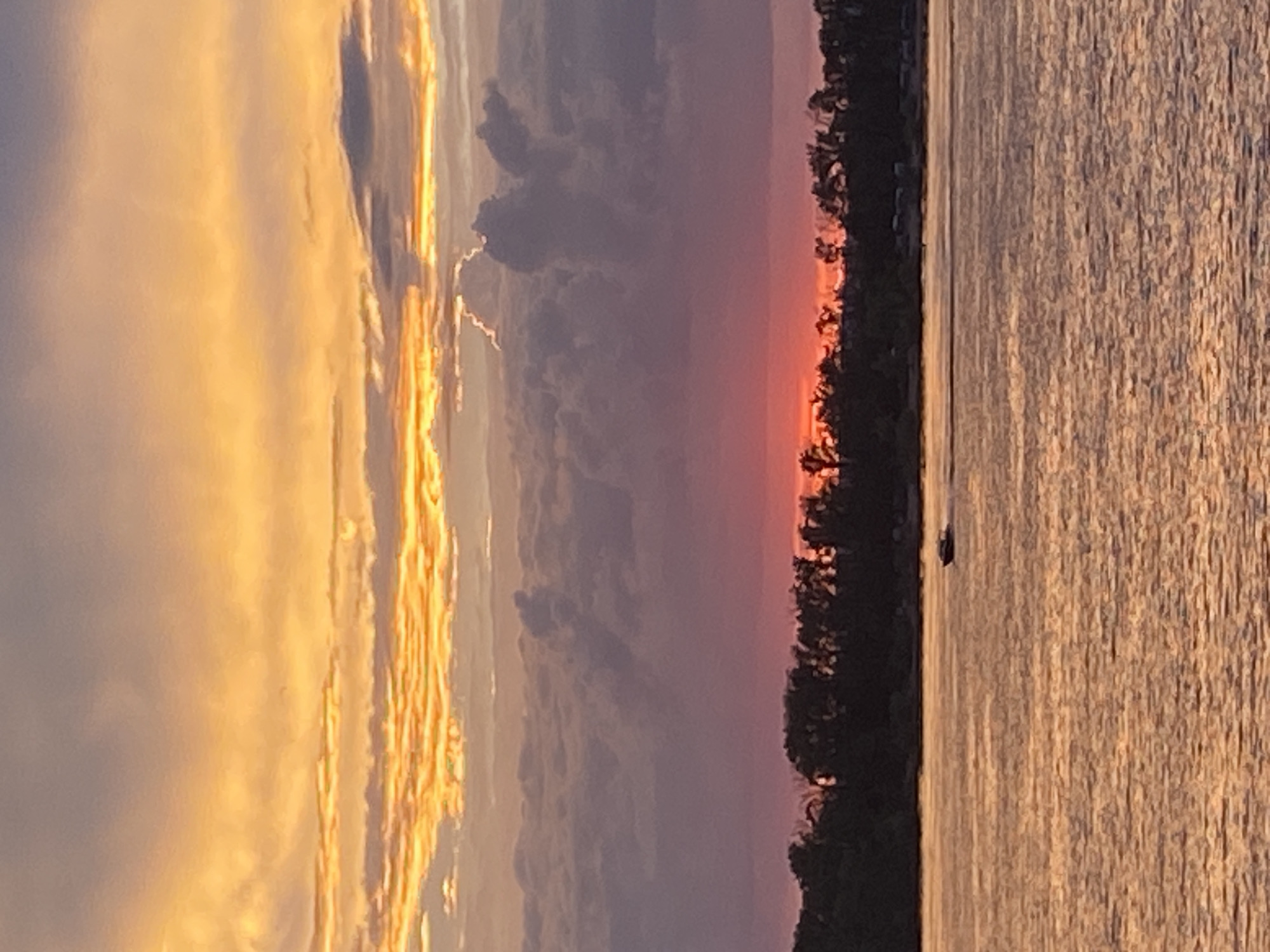 The image captures a sunset over a body of water, possibly the Volga River, near Novokuybyshevsk, Russia. The sky is filled with vibrant colors, transitioning from fiery orange at the top to shades of pink and purple closer to the horizon. Wispy clouds are illuminated by the setting sun, creating a dramatic effect. A dark silhouette of trees lines the distant shore, and a small boat is visible on the water, leaving a faint wake. The water itself reflects the warm hues of the sky, creating a shimmering surface. The scene evokes a sense of tranquility and natural beauty.