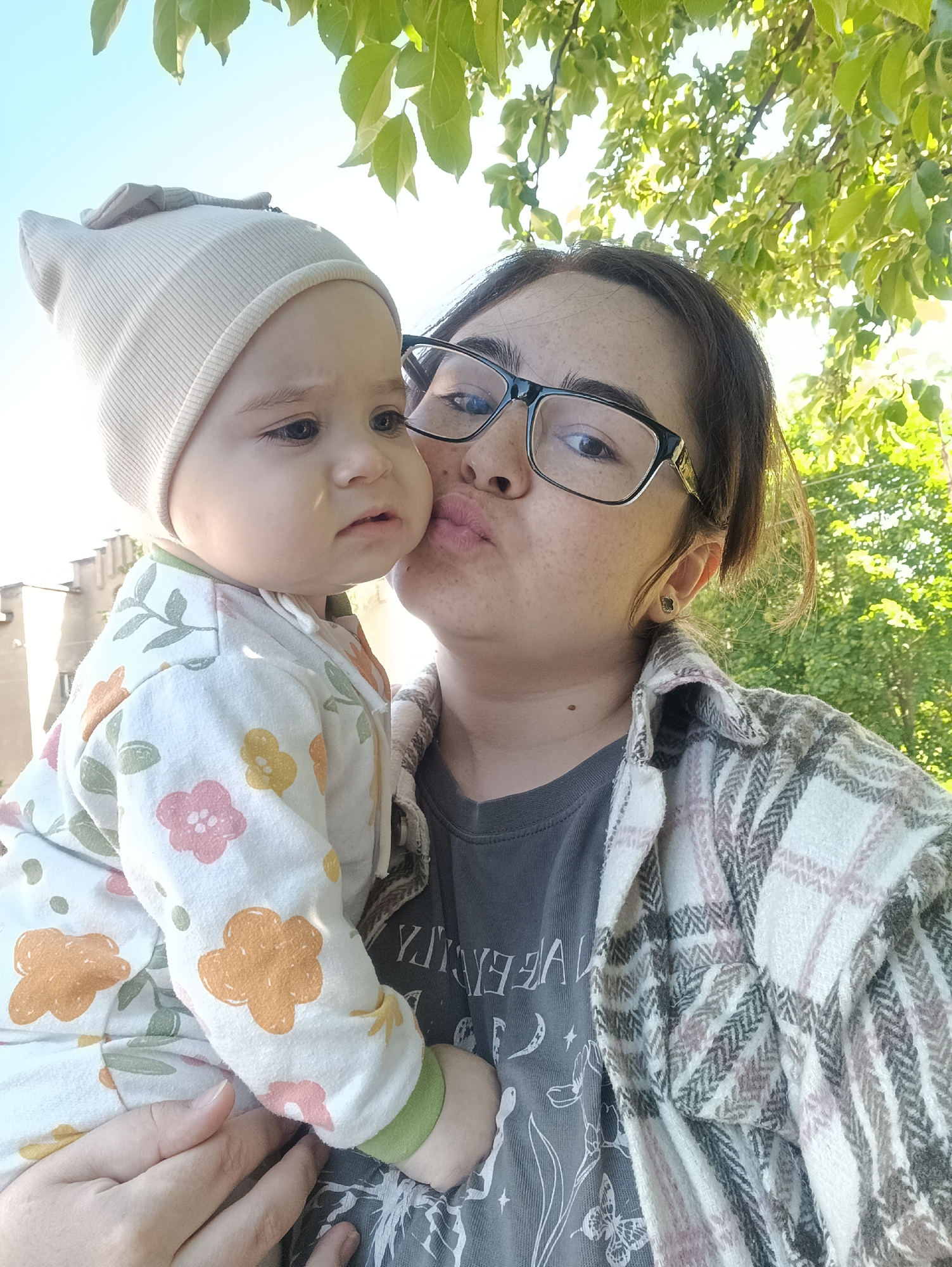 A woman and a baby are captured in a selfie-style shot outdoors under a tree, likely in Noviy, Russia. The woman, who appears to be an adult, wears black-framed glasses, a grey graphic t-shirt, and a white and beige plaid jacket. She is looking towards the baby with her lips puckered, as if giving a kiss or making a playful face. Her dark hair is pulled back, and she has visible freckles.
She is holding a baby, who wears a light-colored ribbed beanie with a small ear-like detail and a white long-sleeved jumpsuit patterned with orange, pink, and green flowers and leaves. The baby is looking away from the camera and the woman, towards the left, with a somewhat serious or slightly pouting expression.
The background shows a bright, clear sky, with lush green leaves from a tree overhead, suggesting a sunny day in an outdoor setting like a park or garden. Vague architectural elements of a building are visible in the distant left background.
On the woman's grey t-shirt, faint white graphics are visible. Part of an inverted word resembling "VALLEY" or "VALLEYR" can be seen, accompanied by illustrations of crescent moons, stars, various plant stems, and a butterfly. The overall mood is one of affectionate interaction between the two subjects in bright daylight.