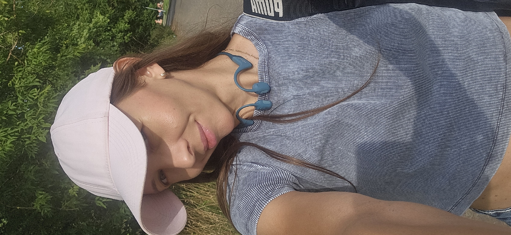 A woman with long brown hair, wearing a pink baseball cap, a striped blue shirt, and bone conduction headphones, smiles at the camera. She is outdoors, with greenery and a paved path visible in the background. The lighting suggests it is daytime. There are no identifiable landmarks or features that indicate the location is Vitsebsk, Belarus.