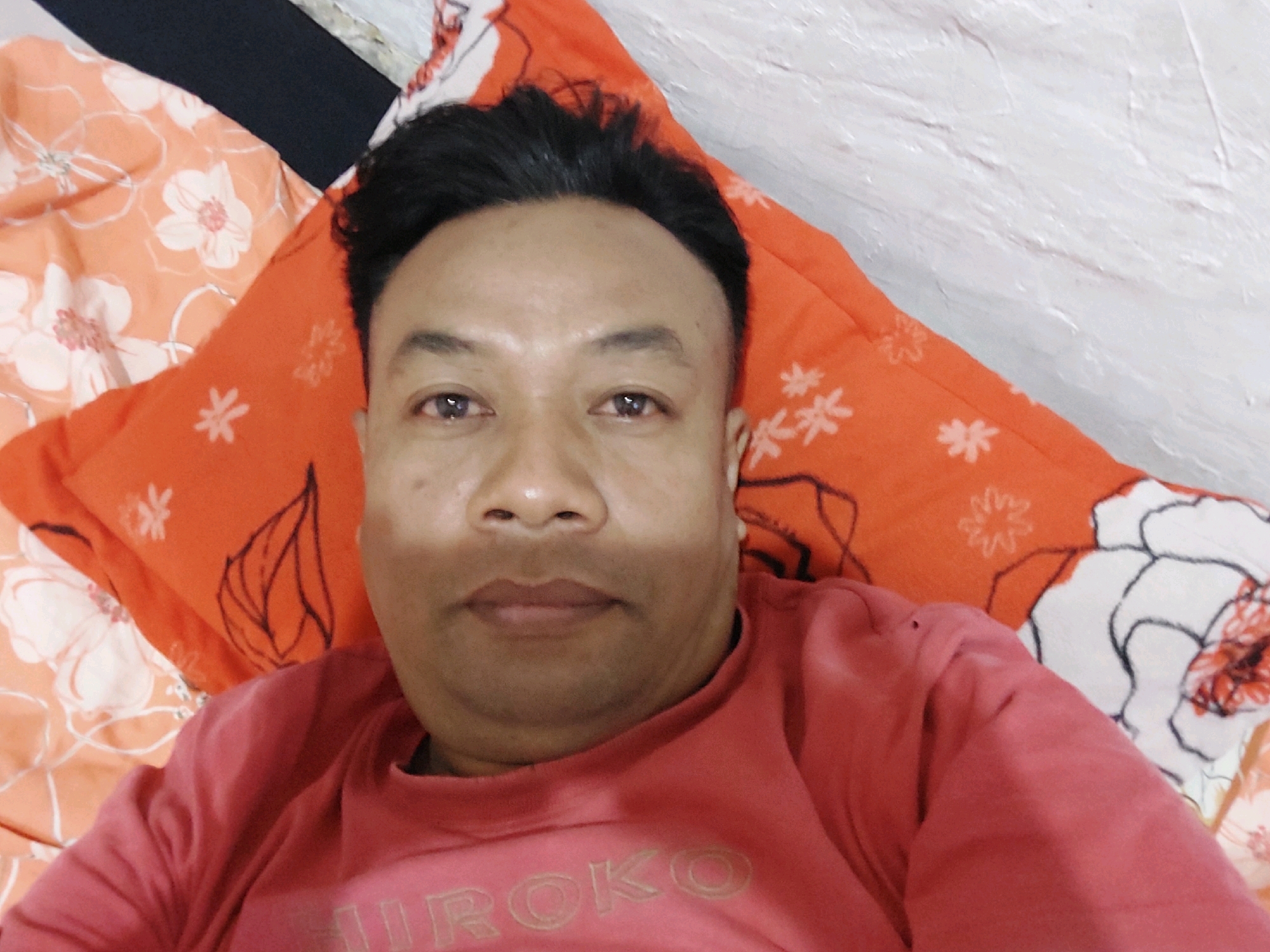 In a close-up, overhead shot, a person with short black hair and brown eyes lies on an orange and white floral patterned surface. The person is wearing a pink crewneck sweatshirt with a white, embossed text that appears to read "HIROKO" across the chest. The background is a textured white wall. The scene is indoors. The location is SARANGAN, Indonesia.
