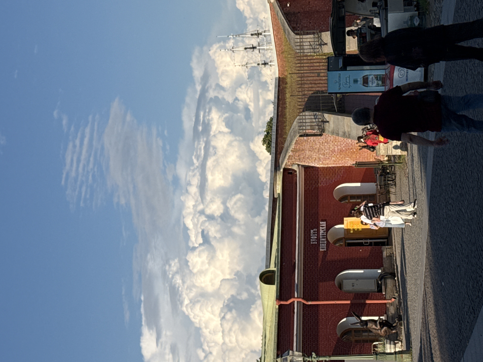 This image depicts a sunny day at the Peter and Paul Fortress in Saint Petersburg, Russia. In the foreground, a cobblestone square is visible with people walking. To the left, a red brick building with arched doorways is prominent, featuring signage that reads "БУФЕТЬ КОНДИТЕРСКАЯ" (Buffet Confectionery). A bronze sculpture of a rabbit stands near one of the doorways. Several individuals are observed interacting in front of the building, and near a food stand to the right, where a bottle advertising "ЧЕРНОПЛОВКА" is displayed. In the background, the sky is filled with dramatic, large cumulus clouds, and the masts of a ship can be seen above the fortress walls. The architecture and historical context suggest this is a tourist area within the fortress complex.