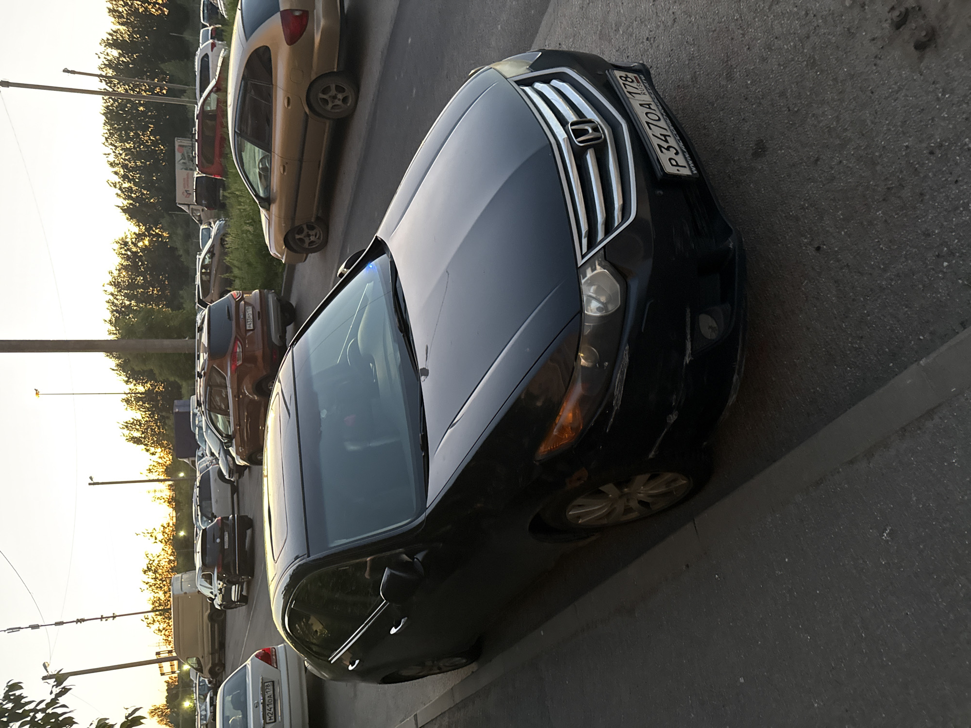 A black Honda sedan is parked on a city street in Saint Petersburg, Russia, in the late afternoon. The car is positioned at an angle, with its front facing the viewer. The license plate reads "P347OA 178". In the background, other vehicles, including SUVs and a gold station wagon, are parked or driving. A line of trees and vegetation runs along the right side of the street, and tall poles with lights are visible in the distance. The sky is illuminated by the setting sun, casting a warm glow over the scene.