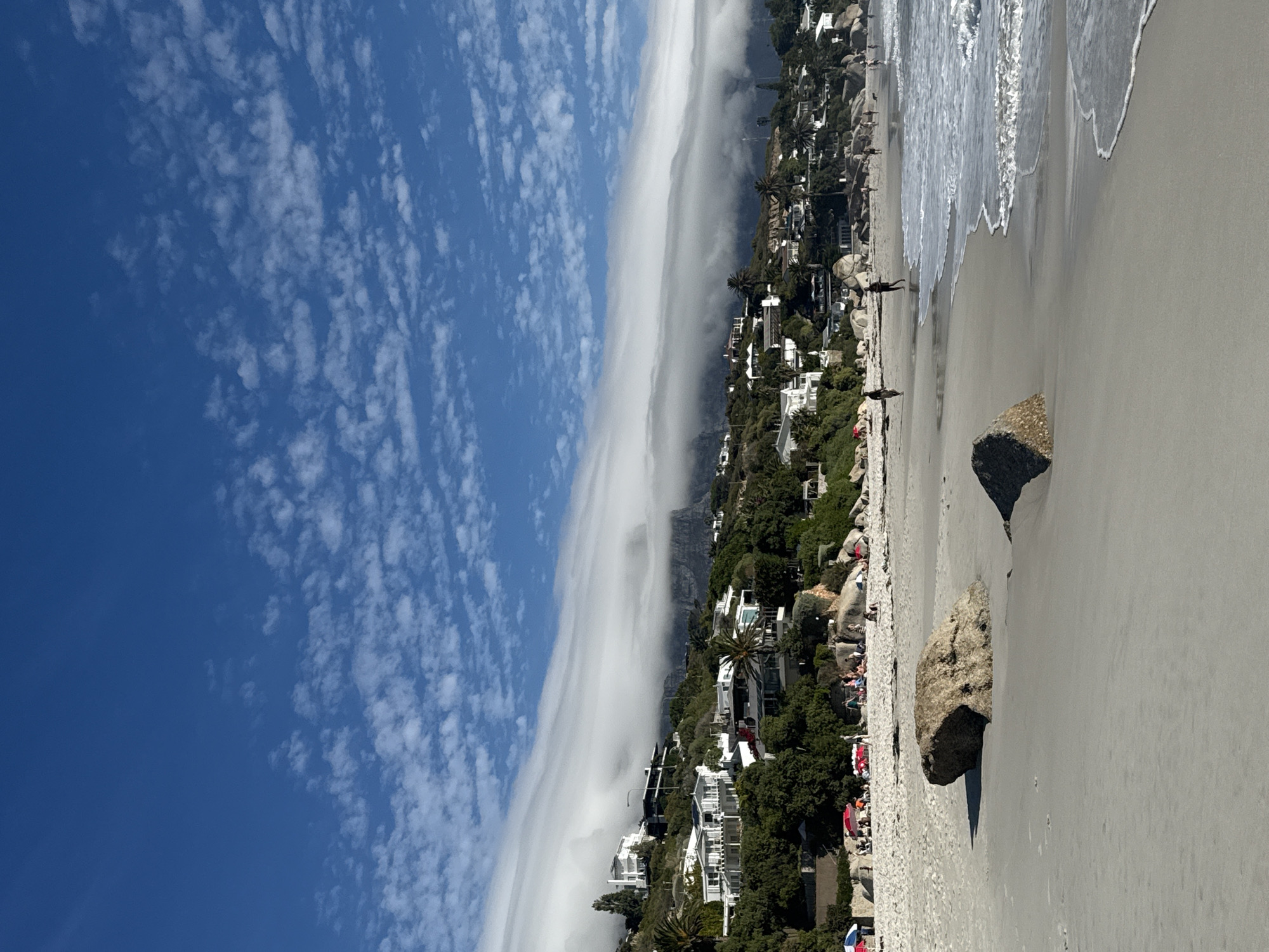The image depicts a daytime coastal scene in Cape Town, South Africa, likely in the Clifton or Camps Bay area, with Table Mountain in the background. The foreground features a wide expanse of light-colored sand with two prominent, dark-brown, weathered rocks. To the right, the Atlantic Ocean meets the shore with gentle white waves breaking.
In the midground, numerous individuals are present on the sandy beach. Several groups are visible, some gathered under colorful beach umbrellas, including red and blue. Other individuals are walking along the wet sand closer to the waterline. At least five distinct human figures are observable in this area, engaged in leisure activities.
The background is dominated by a lush, green hillside rising directly from the beach, densely populated with modern, multi-story white residential buildings featuring flat roofs and balconies. These structures are interspersed with mature trees and palm trees. Above this residential area, a thick, horizontal cloud formation, characteristic of the "Tablecloth," drapes over the upper portion of a large mountain range, partially obscuring its peak. The sky above is a clear, vibrant blue with scattered, small white cumulus clouds.