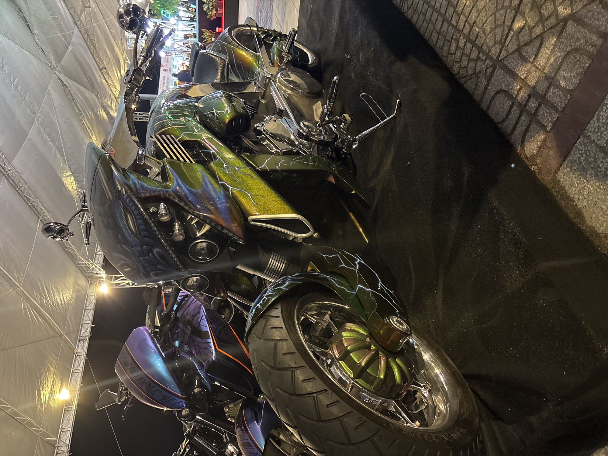 The image shows two custom motorcycles displayed on a dark cloth covering a sidewalk in Nha Trang Ward, Vietnam. The sidewalk is paved with a patterned concrete design. The motorcycle in the foreground has a green and black paint job with lightning bolt designs. The front wheel has a custom hubcap with a geometric pattern. The motorcycle behind it has a purple and black paint job. The background shows a tent structure with lights. In the distance, there are other motorcycles, people, and storefronts. The lighting suggests it is evening.