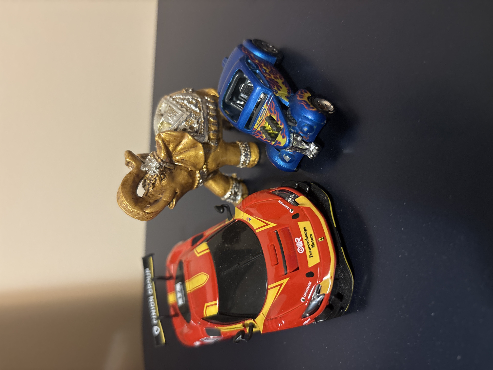 The image shows a gold-colored elephant figurine, a blue toy car with flame decals, and a red and yellow toy race car with "Francorchamps Motors" and "Orifarm" printed on it. They are arranged on a dark blue surface next to a light beige wall. There is no indication of this being Plovdiv, Bulgaria.