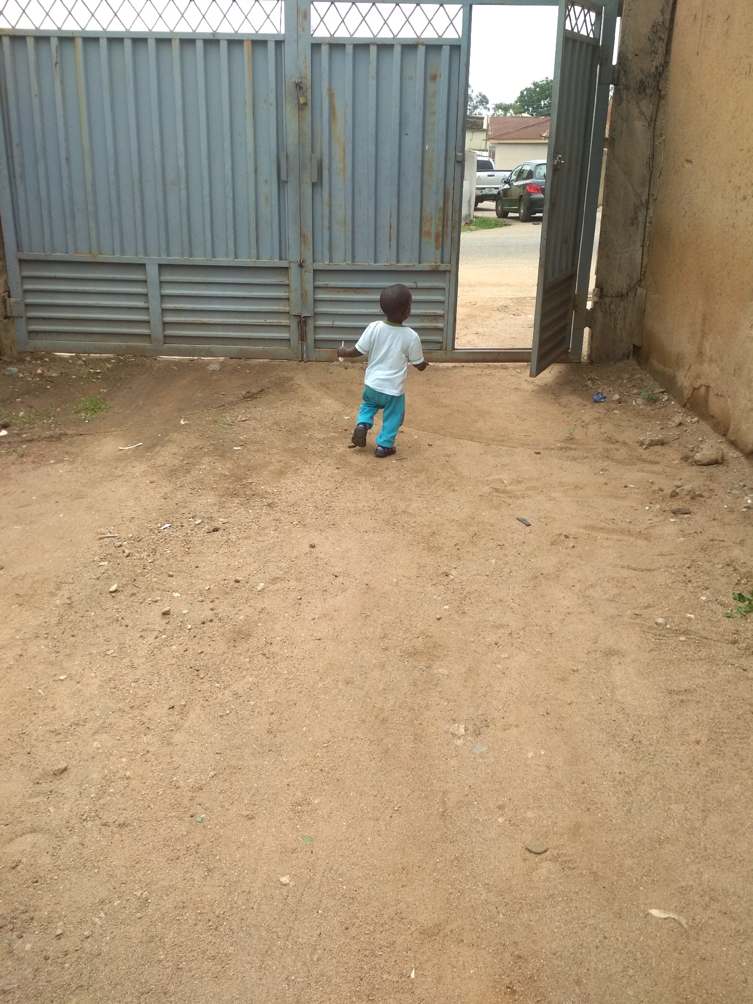 A small child in a white shirt and blue pants is walking out of an open gate onto a dirt yard. The gate is gray and made of metal, and the yard is sparsely covered with dirt and small pieces of debris. The right side of the scene features a portion of a wall that is brown and aged. In the background, outside the gate, there are cars parked on a street. The location is in the city of Unknown, Unknown.