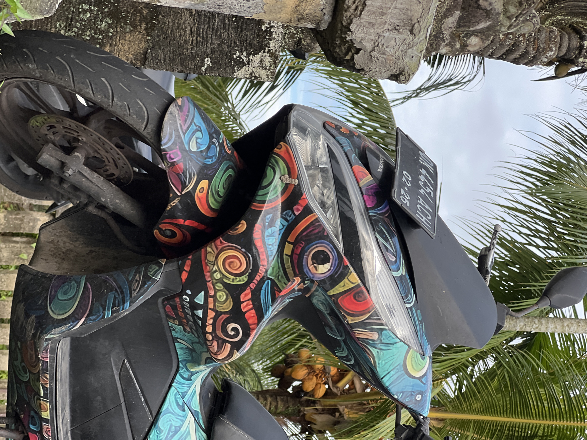 The image depicts a motorbike in Ubud District, Indonesia. The motorbike is adorned with a colorful, swirling pattern on its body. A license plate is visible. The background includes palm trees, suggesting a tropical environment. The weather appears overcast.