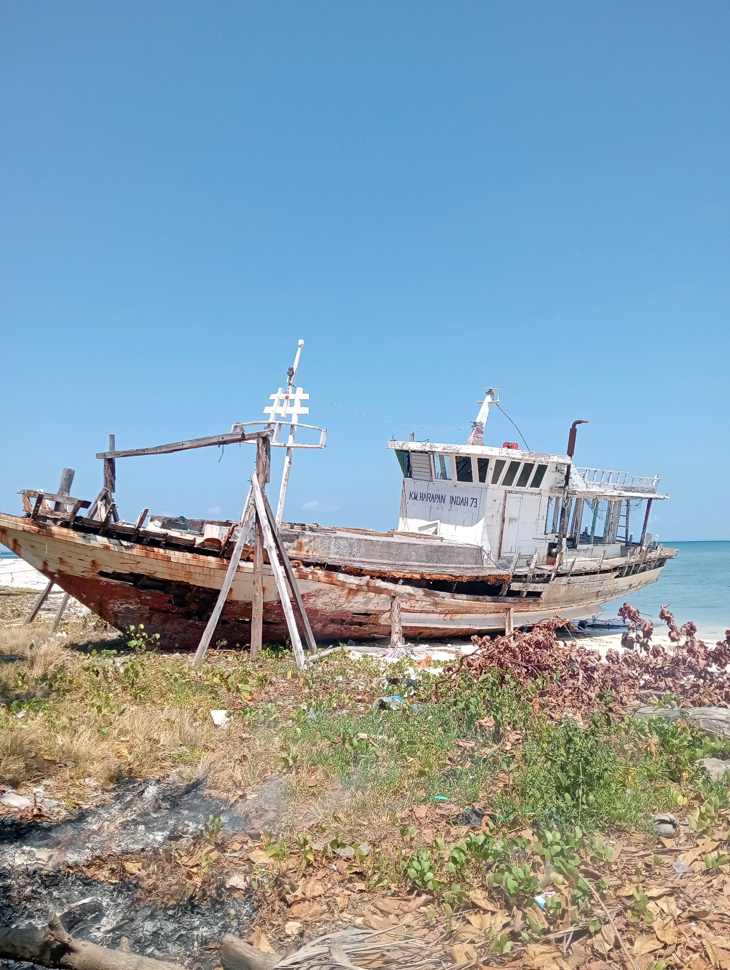 A dilapidated wooden ship, identified by the inscription "KM HARAPAN INDAH 73", rests aground on a sandy beach under a clear blue sky. The ship, showing significant signs of decay and rust, is supported by wooden scaffolding. The surrounding environment is a mix of dry grass, fallen leaves, and sparse green vegetation. In the background, the calm blue ocean meets the horizon. The scene suggests a location in Pagerungan Kecil, Indonesia, specifically a coastal area where abandoned vessels might be found. There are no people visible in the image, and no explicit actions or interactions are depicted.