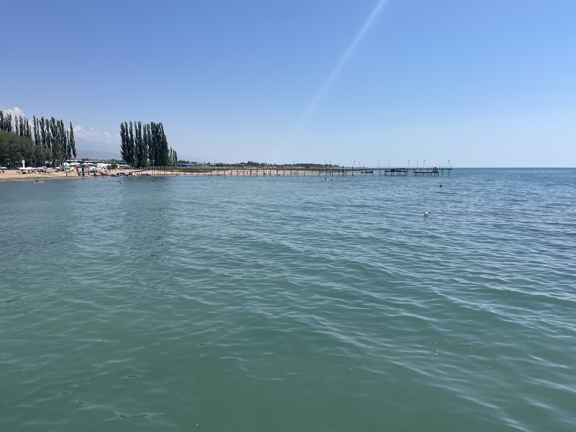 The image depicts a serene view of a body of water on a clear day, likely in Ornek, Kyrgyzstan. The foreground is dominated by the rippling, turquoise surface of the water. In the mid-ground, a sandy beach is visible, dotted with numerous colorful umbrellas and beachgoers enjoying the sun. Several tall, slender trees line the edge of the beach, creating a distinct visual boundary. Beyond the beach, a wooden pier extends out into the water, with a few people visible on it. The horizon is a sharp line separating the calm water from a vast, clear blue sky. The overall atmosphere suggests a tranquil summer day at a popular lakeside or seaside resort.