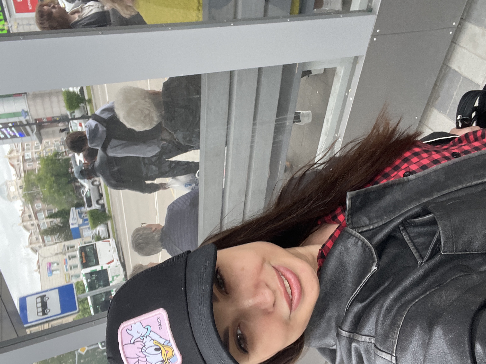 A woman in a black cap with a "Daisy" patch, a red and black plaid shirt, and a black leather jacket is taking a selfie at a bus stop. The bus stop is in an urban area with buildings, buses, and street signs visible in the reflection of the bus stop shelter. Several people are reflected in the glass, including a woman with blonde hair, a man in a gray shirt, and a group of people walking down the street.