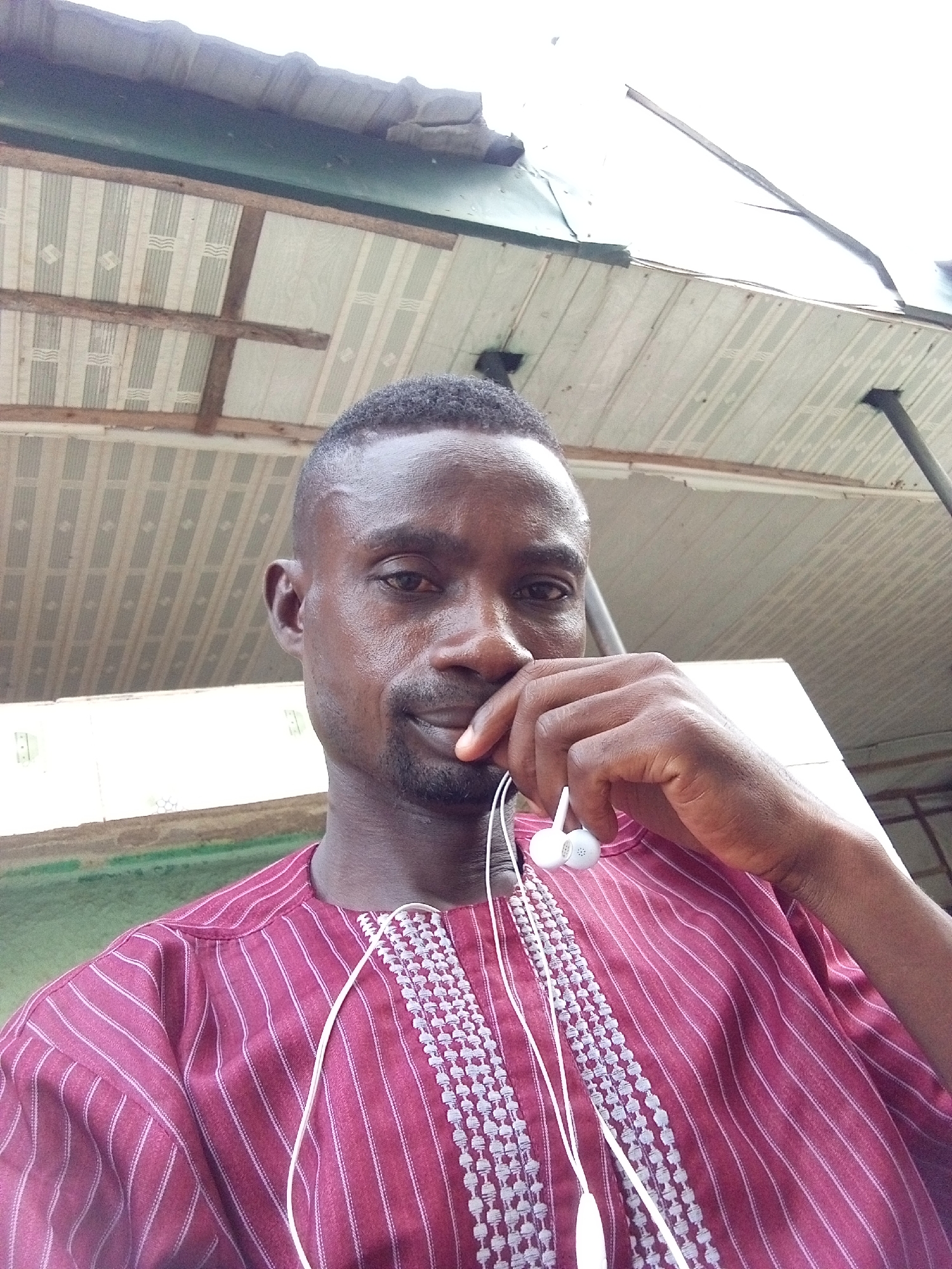 A close-up selfie shows a man with dark skin, short hair, and a trimmed beard looking directly at the camera. He is positioned slightly below the camera, gazing upwards into the lens. His right hand is raised, with his thumb resting against his lower lip, suggesting a thoughtful or slightly reserved pose. He wears a maroon or burgundy long-sleeved shirt with subtle white vertical stripes. The shirt features an ornate, light-colored embroidered placket down the front, consistent with traditional attire often seen in Nigeria. White in-ear headphones hang around his neck, their wires draping over his chest and near his hand.
The background reveals an overhead structure, likely a ceiling or canopy. It consists of panels with a light striped pattern, possibly in white and light green, supported by visible wooden beams. Above this, a corrugated metal roof is partially visible, suggesting an outdoor or semi-outdoor setting under shelter. The lighting is bright and diffuse, indicative of daytime, possibly under an overcast sky. The overall impression is a casual, personal shot taken in Zango-ndaji, Nigeria, with the man's expression being neutral to slightly contemplative. There is no visible text in the image.