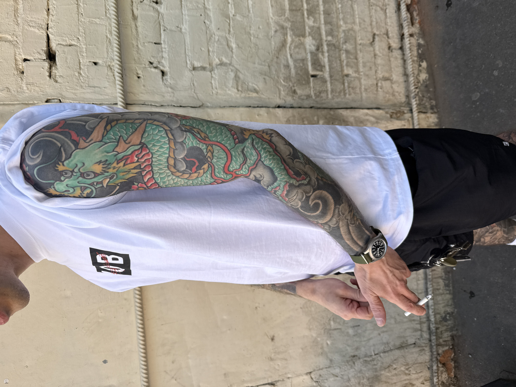 An individual, identified as male, is depicted standing outdoors, possibly in an alley or a side street in Moscow, Russia. The subject is shown from the chest down, facing slightly left of the viewer, with his left arm brought forward. He wears a white sleeveless t-shirt featuring a black rectangular logo on the chest with the letters "OB" crossed by a red line, and black shorts. His right arm is covered by a full-sleeve Japanese-style tattoo of a vibrant green and red dragon against a dark, cloud-like background. The left arm also has a dark-toned sleeve tattoo, partially obscured by the t-shirt. On his left wrist, he wears a military-style watch with a green fabric strap and a dark face. He holds a lit cigarette between the index and middle fingers of his left hand. A set of keys is clipped to his black shorts on the left hip. The background comprises two distinct walls: an older, textured light beige wall on the left with a horizontal decorative molding, and a light beige brick wall on the right with visible cracks and wear. The ground is dark asphalt or concrete. The scene is illuminated by natural daylight.