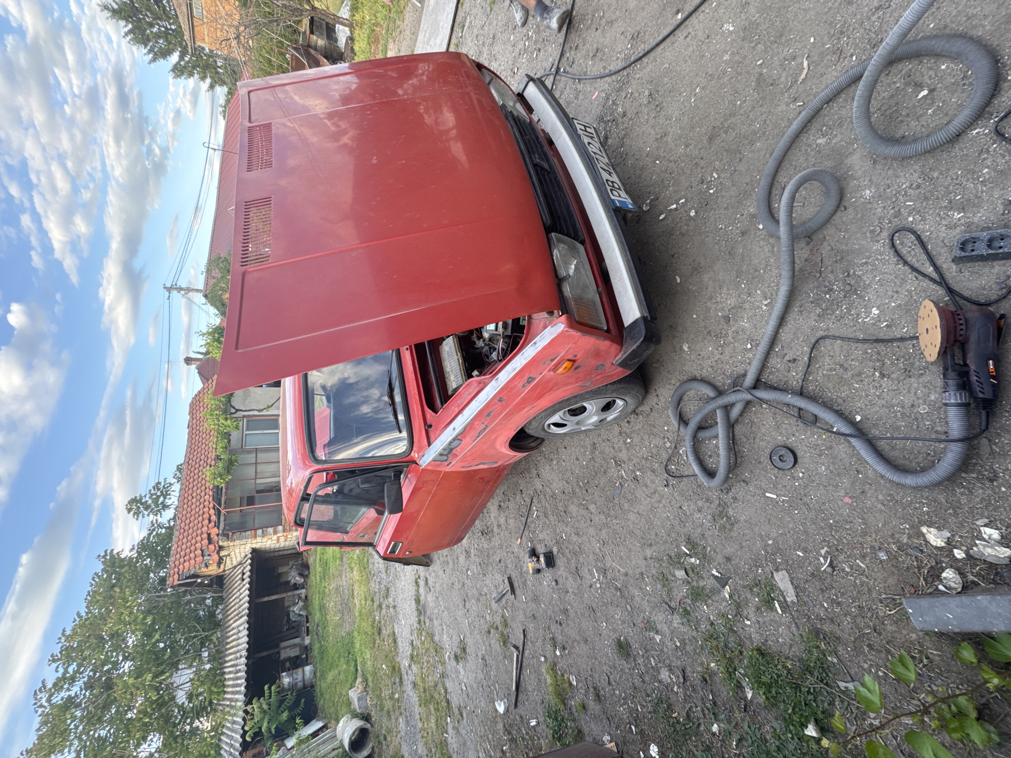 The image shows a red Lada car with its hood open, parked on a dirt and gravel surface. The car is likely undergoing repairs or maintenance. To the right of the car are tools including a power sander, a gray hose, and an electrical power strip. In the background, a weathered building with a damaged roof is visible among trees, and a cloudy sky overhead. The license plate number is visible as PB 4712 AH. The location is Radinovo, Bulgaria.