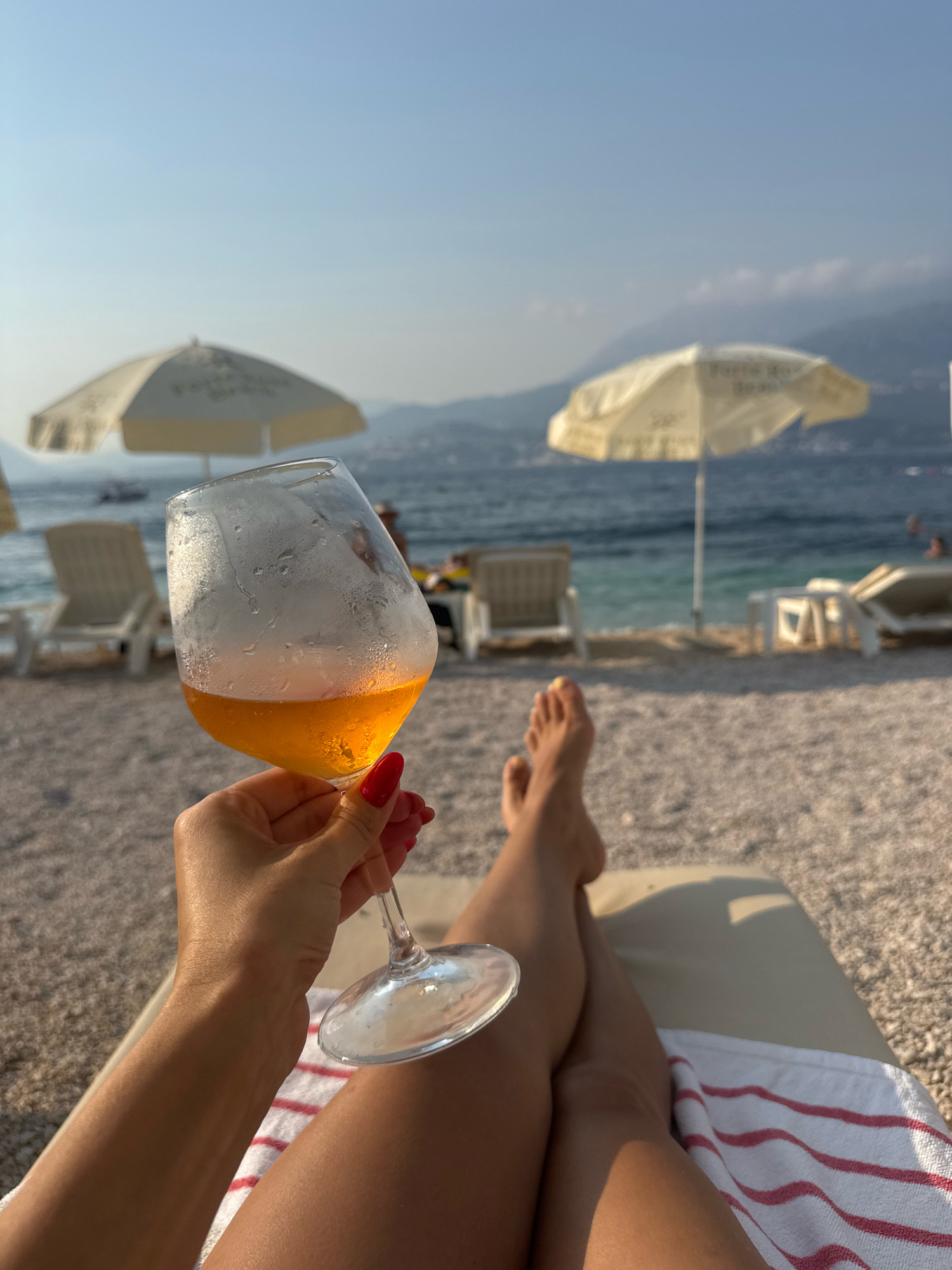 In an outdoor, beachfront setting, a person's hand with red nail polish holds a stemmed wine glass filled with an orange beverage. The glass is partially covered in condensation. The person is lying on a lounge chair with a striped towel draped over their legs. Their feet are visible, and they are wearing sandals. In the background, the sea is visible, with mountains and a clear sky. There are several beach umbrellas and lounge chairs on the pebbled beach.