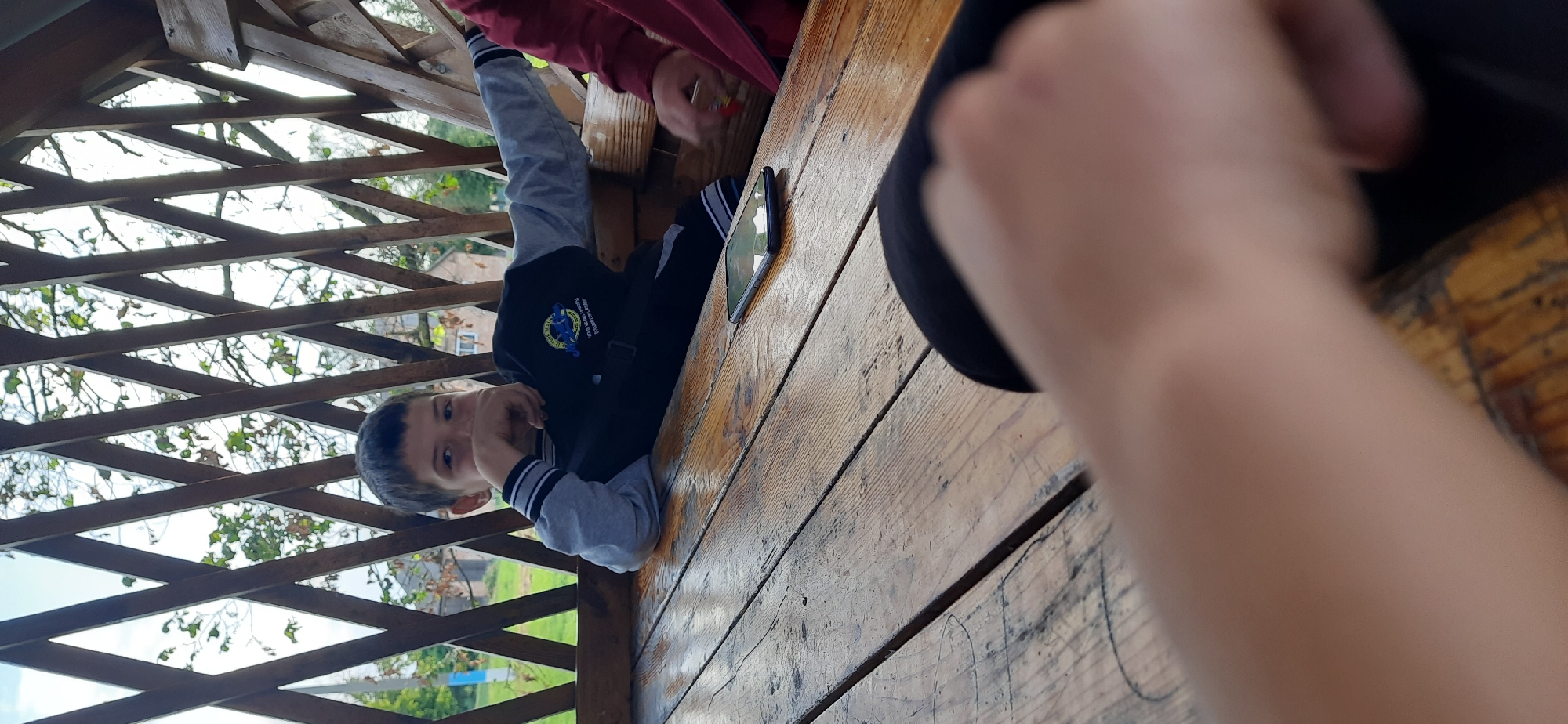 In the foreground, a young boy lies on a wooden picnic table, his body angled away from the viewer. He wears a black and gray jacket with white stripes on the cuffs. His face is partially visible, turned towards the camera with an inquisitive expression. A smartphone rests on the table next to him, its screen displaying a colorful image.
The scene is set outdoors, likely within a gazebo or similar structure, indicated by the wooden beams and latticework overhead, through which dappled sunlight filters. Trees and foliage are visible in the background, suggesting an outdoor natural setting. The light suggests it is daytime. The mood appears casual and relaxed. There is no discernible text in the image.