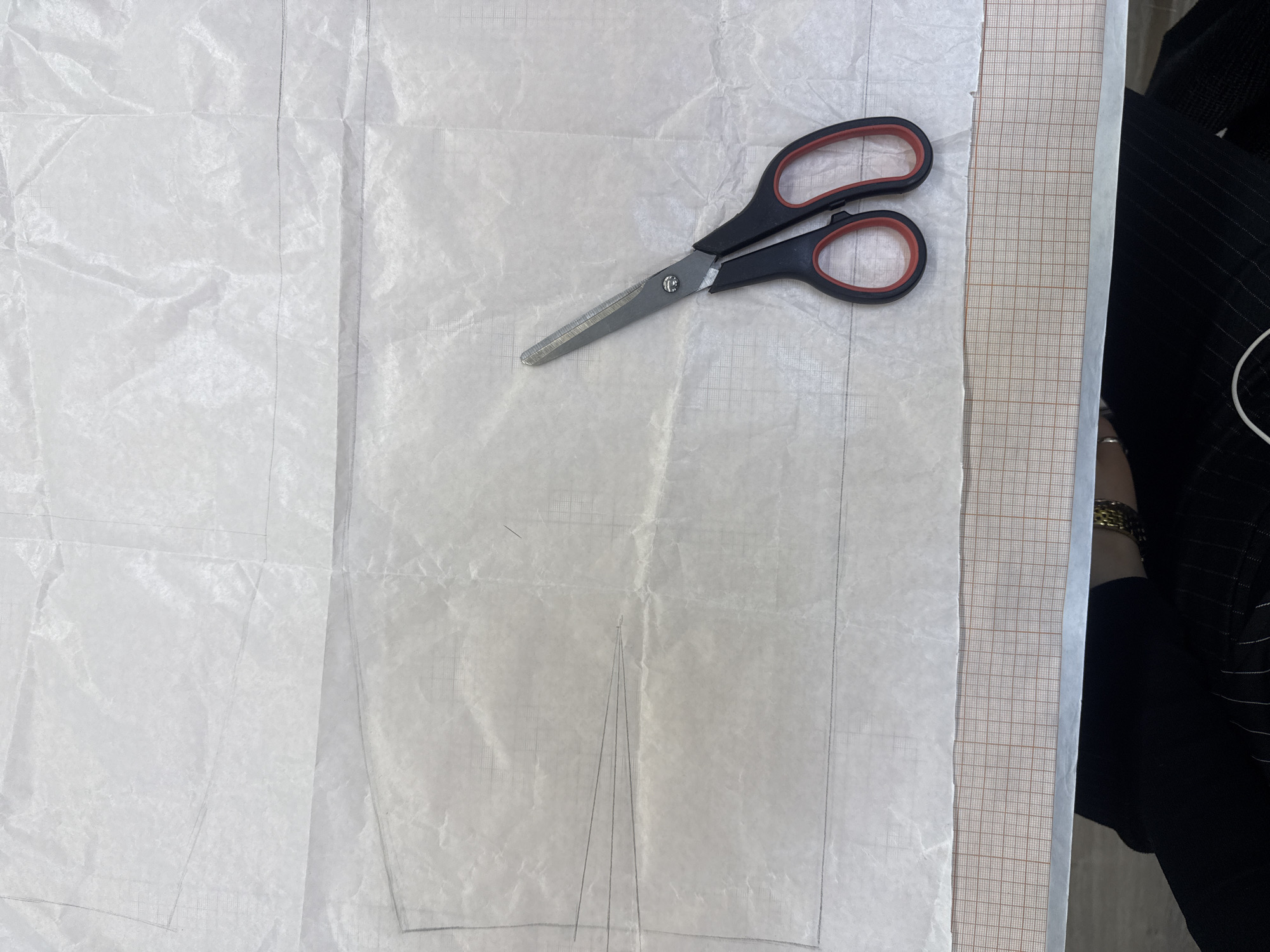 The image shows a pair of scissors lying on a sheet of paper. The paper appears to have a sewing pattern drawn on it in pencil. The paper lies on a grid-patterned surface. A person's arm with gold-colored bracelets is visible in the upper right corner, wearing a black and white striped garment. The location is not discernible from the image.