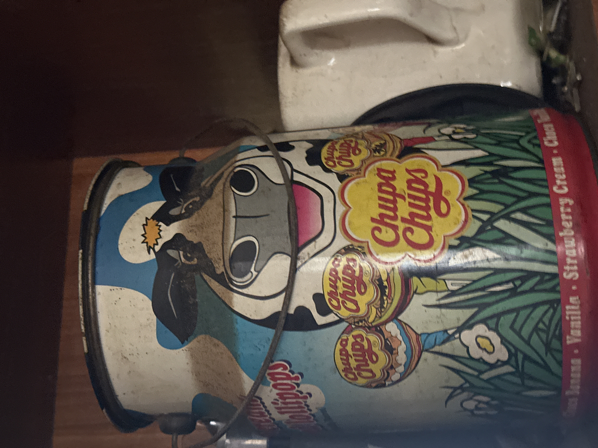 The scene, located in Juan Griego, Venezuela, depicts an indoor setting focused on a vintage-style cylindrical metal container, approximately 30-40 cm in height, resembling a milk can. The container displays a printed design featuring a stylized cartoon cow's head with black, white, and grey tones, pink accents on its mouth, and black eyes. A yellow starburst graphic is positioned above the cow's head. The upper portion of the container shows abstract blue and white cloud-like patterns. Below the cow, three distinct Chupa Chups lollipop logos are prominent: two smaller ones on the left with various wrapper designs, and one larger, central logo. This section is set against a light blue backdrop with green stylized grass and white flowers at the container's base. The bottom rim of the container is a red band with partially legible white text, identifying flavors such as "Banana," "Vanilla," "Strawberry Cream," and "Choco." A dark, thin wire handle is symmetrically attached to the upper sides of the container. The container's surface exhibits signs of age, including accumulated dust, scuffs, and minor paint wear. To the right of the container, a partial view of a white ceramic object, possibly a pitcher handle, is visible. The immediate background behind the container consists of a dark brown, indistinct surface, likely wood, suggesting a shelf or cabinet interior. The surface directly beneath the container is dark and shows minimal debris. No people are visible.