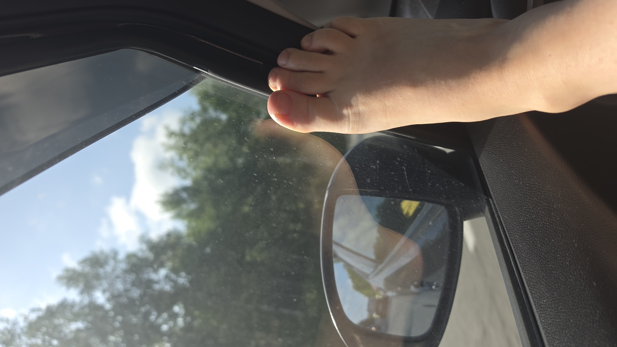 In an exterior shot of a car interior, a bare foot is visible, propped on the car door frame near the side mirror. The foot is light-skinned with visible toes and toenails. The side mirror reflects a blurry image of a car and trees. Outside the car, the sky is blue with scattered clouds, and trees are visible through the window. The interior of the car appears to be made of black textured plastic. The image is taken during the day, with sunlight creating shadows and highlights. The specific location in Kruglikovo, Russia is not discernible from the visual information provided.