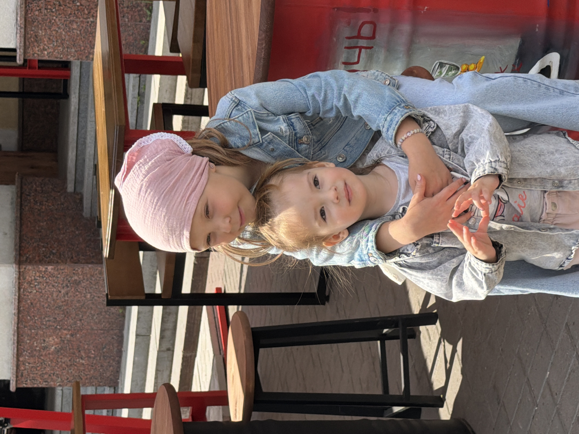 Two young girls are posing for a photograph outdoors on a sunny day, in the city of Ufa, Russia. The older girl, on the right, wears a pink headscarf and a denim jacket. She has her arm around the shoulders of the younger girl, who is on the left and faces the camera. The younger girl wears a denim jacket over a t-shirt and light-colored pants. They are situated in front of a red cylindrical object with some Cyrillic writing and an image painted on it. In the background, there are outdoor tables and chairs, with a stone building and steps behind them. The ground is paved with brick-like stones. The scene appears to be in a cafe or restaurant outdoor seating area.