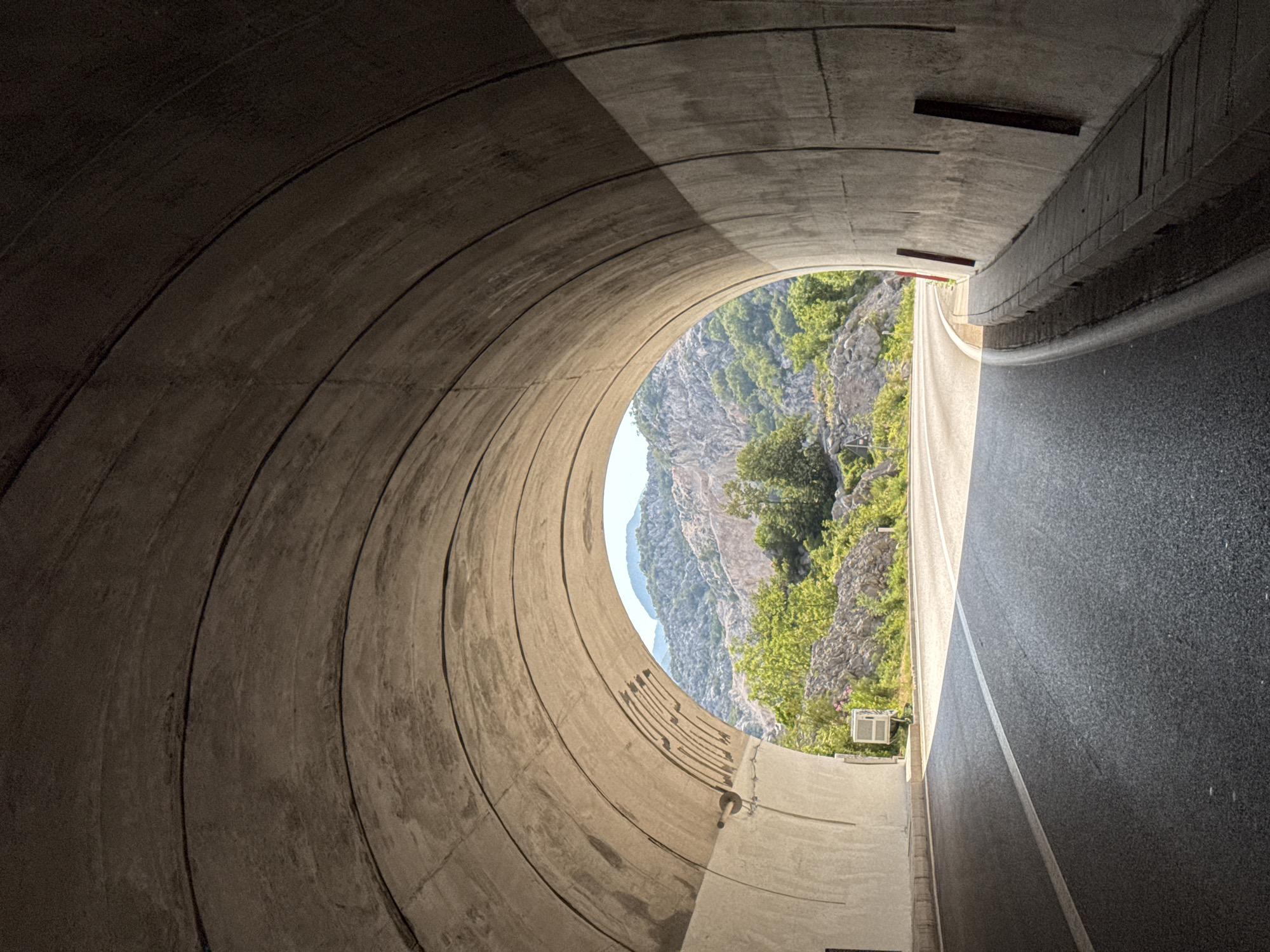 The image shows the exit of a concrete tunnel on a sunny day. The tunnel has a curved ceiling with visible expansion joints. A paved road curves out of the tunnel and into a mountainous landscape with green trees and rocky terrain. The road has a white line marking the lane. On the left side of the tunnel exit, a small white air conditioning unit is visible. On the right side, there is a concrete curb and what appears to be a drainage channel. The overall scene suggests an infrastructure element integrated into a natural environment. The location is the Risan tunnel on the coast of Kotor Bay, Montenegro.