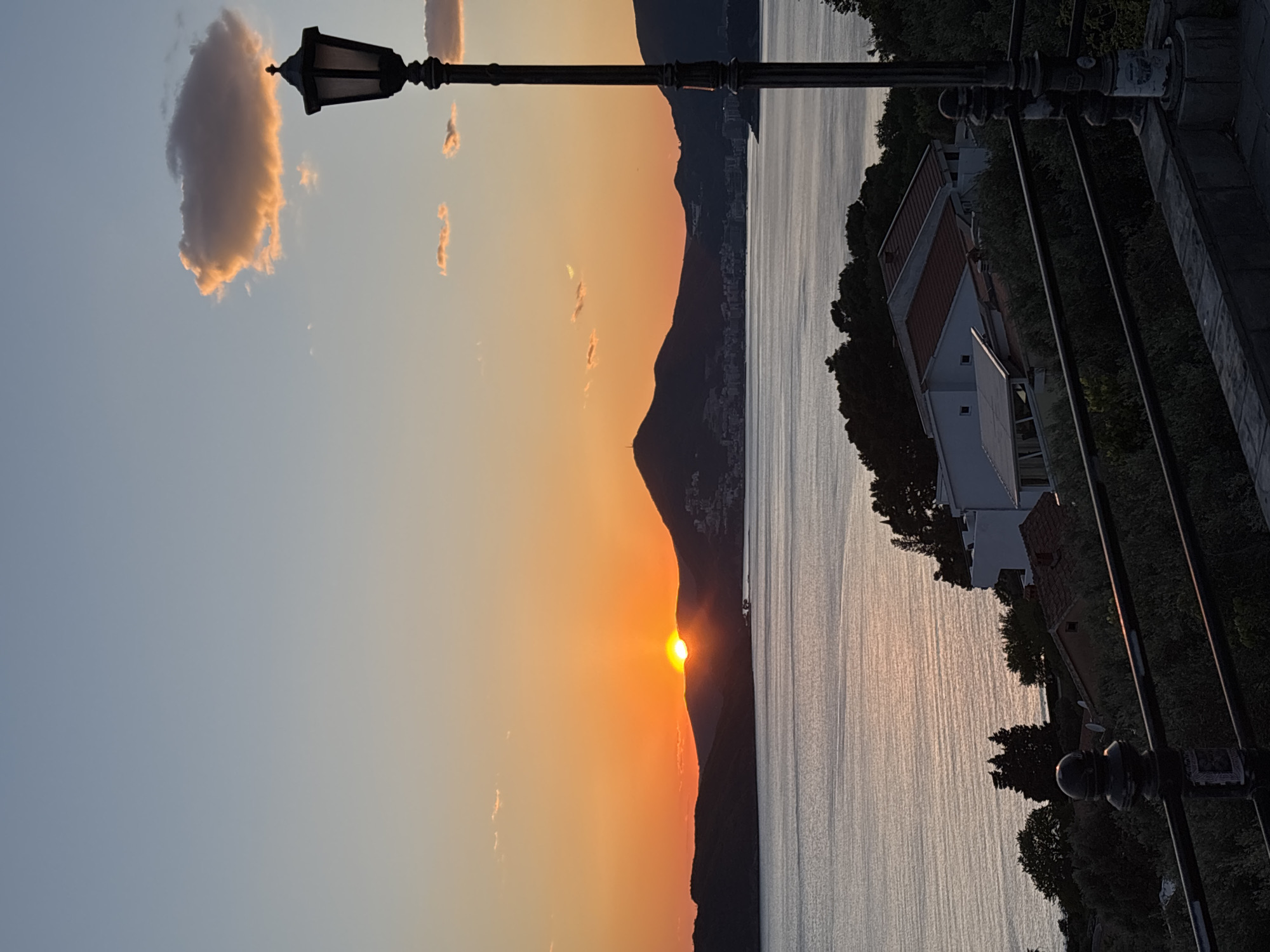 The image depicts a sunset over the water, likely a sea or a large lake. A mountain range silhouette is visible in the distance, with the sun setting just behind it. In the foreground, there is a black lamppost extending into the frame. A building or structure with a reddish roof is situated on the right side of the image, next to trees and greenery. The sky transitions from a light blue at the top left to a vibrant orange near the horizon where the sun is setting. A few scattered clouds are present in the sky. The water reflects the colors of the sunset. The scene appears to be taken from a slightly elevated position, possibly a walkway or a balcony, as a metal railing is visible in the lower right.