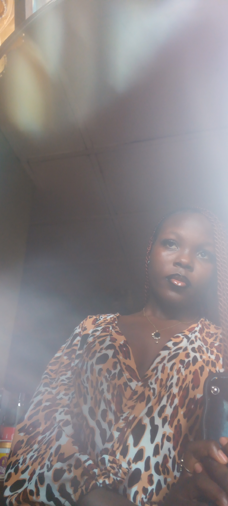 A medium shot captures a young Black woman looking upwards and slightly to her right, her head and shoulders filling the frame. She has dark skin and her hair is styled in intricate, reddish-brown braids, one of which is visible on the right side of her head. She wears a V-neck top with a bold, all-over leopard print pattern in shades of brown, black, and white. A delicate gold necklace with a dark pendant rests on her chest. Her lips are painted with a dark, glossy lipstick, and she appears to be wearing eye makeup. Her expression is somewhat neutral, with a hint of a thoughtful gaze.
The lighting in the image is soft and diffused, with bright highlights creating a hazy, almost ethereal effect, particularly on the left side of the frame and over the subject's face and upper body. This suggests a bright light source, possibly from a window or overhead lighting, creating lens flare and glare. The background is dimly lit and indistinct, appearing to be an indoor setting with a ceiling visible at the top of the frame, marked by intersecting lines that suggest ceiling tiles or panels. The overall atmosphere is somewhat intimate, perhaps a selfie taken in a domestic or personal space. There is no discernible text visible in the image.