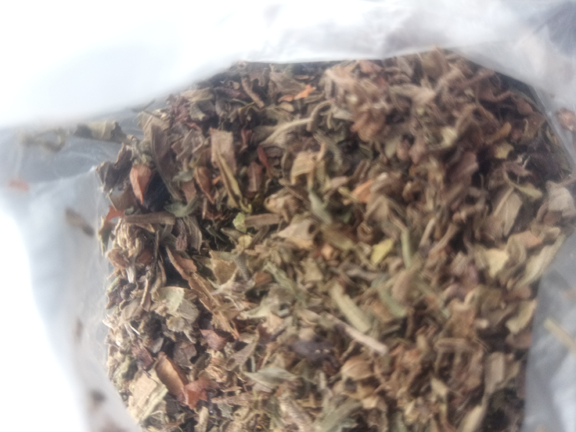 The image shows a close-up, blurry view of dried herbs or plant matter inside a clear plastic bag. The material appears to be a mix of various dried leaves and stems, with colors ranging from light brown and tan to green and some reddish-brown hues. Some pieces are fragmented, while others are more intact. The lighting is bright, causing some overexposure, particularly in the upper left portion of the image, which appears to be a white, possibly out-of-focus background or reflection. The texture of the plant matter is rough and irregular. The image lacks any discernible geographical context of a city or specific location.