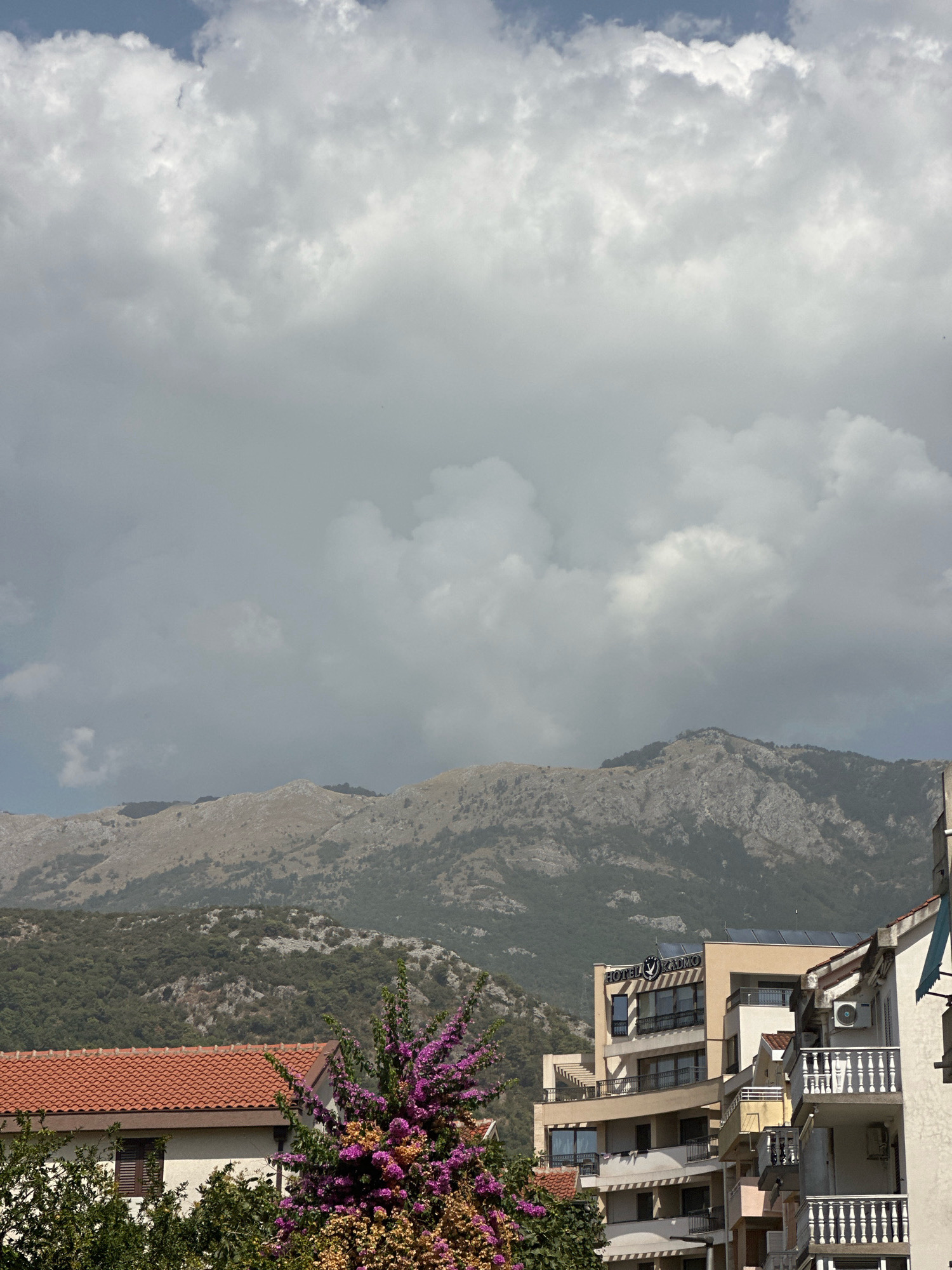 The image shows a view of buildings and mountains under a cloudy sky. In the foreground, there are several buildings with red tile roofs and balconies. One building has a sign that reads "HOTEL RADMO". There is also a bush with purple flowers. In the background, there are mountains covered with trees. The sky is filled with large, white clouds. The scene appears to be in Budva, Montenegro, given the architectural style and the mountainous landscape.