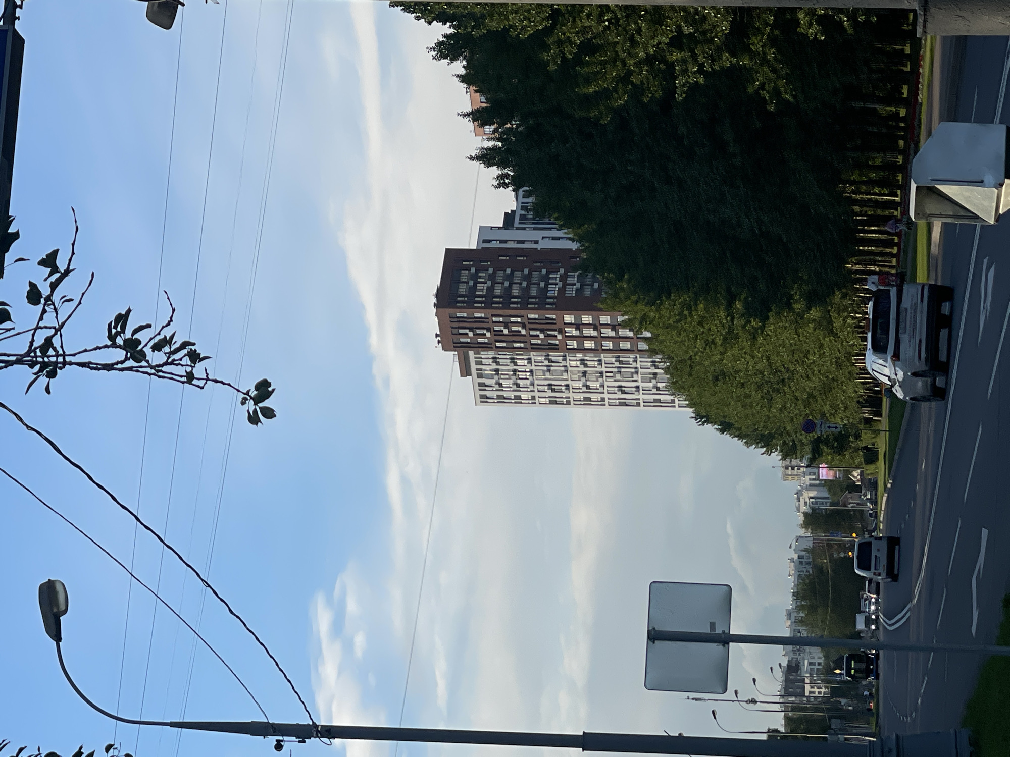 The image captures a street scene in Moscow, Russia, with tall residential buildings lining the right side of a road. A dense line of lush green trees separates the buildings from the road. The sky is a bright blue with scattered white clouds, suggesting it is daytime, likely late morning or early afternoon. The sun is casting shadows, indicating a bright, possibly warm day.
On the road, several cars are visible, suggesting traffic is flowing. One white van is particularly noticeable, driving on the road. A light-colored utility box or structure stands by the side of the road on the right.
In the foreground and to the left, a street light is visible, and thin branches of a tree with sparse leaves hang down. Overhead power lines crisscross the sky. The buildings in the background are a mix of modern, multi-story structures with many windows, appearing to be apartment complexes. One prominent building is dark brown with a grid of white-framed windows. Further in the distance, more buildings are visible, some with lighter facades.
A blank, rectangular sign is mounted on a pole in the middle ground, facing the road. The overall scene depicts a typical urban environment, likely a residential area with active transportation. There is no visible text in the image.